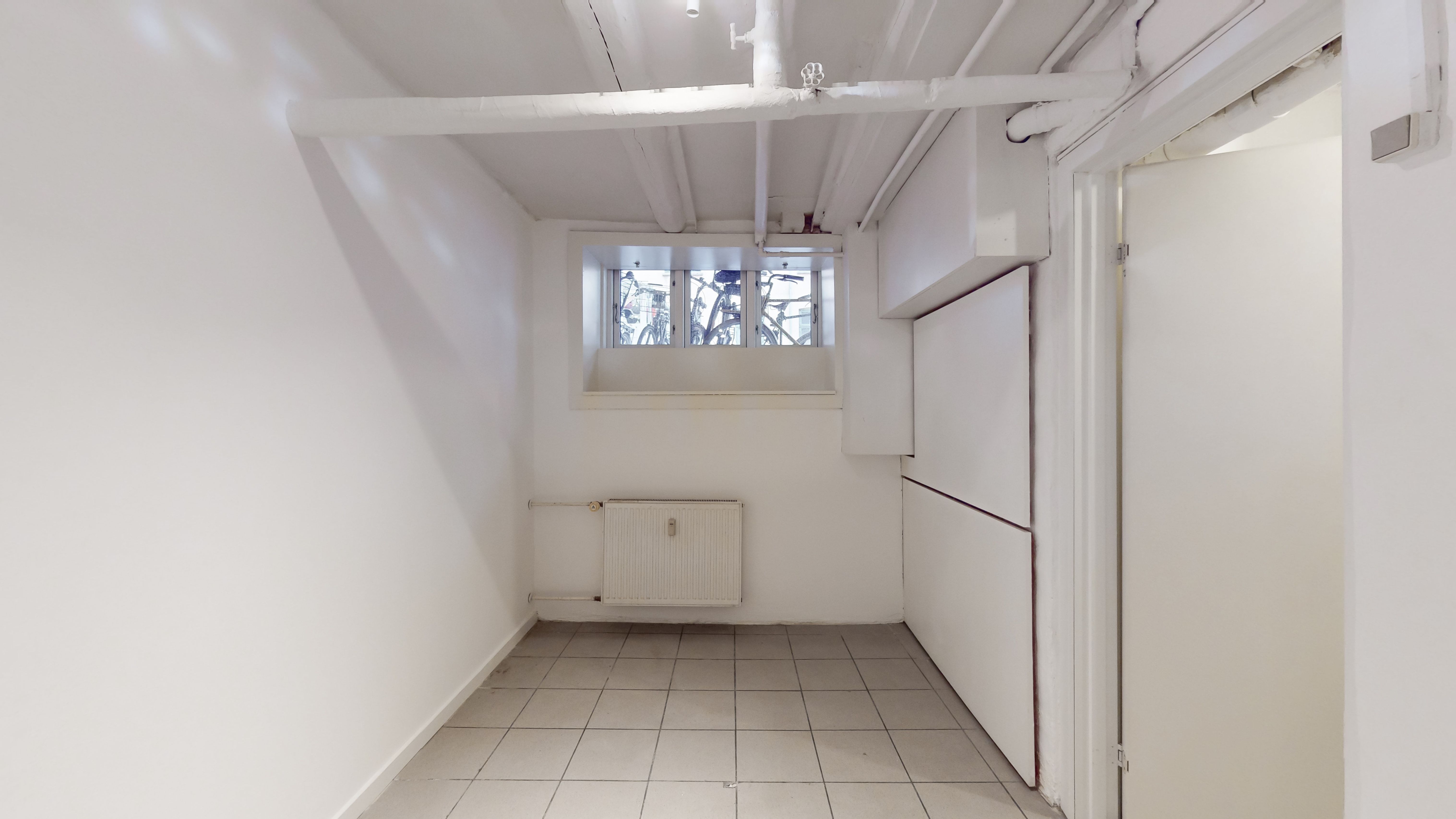 Butikslokale med hvidt loft og vægge, hvor der er et vindue med udsigt til gaden. Der er en hvid radiator og et skarpt, hvidt gulv.