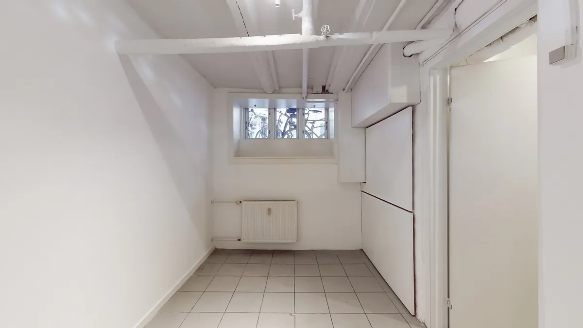 Butikslokale med hvidt loft og vægge, hvor der er et vindue med udsigt til gaden. Der er en hvid radiator og et skarpt, hvidt gulv.