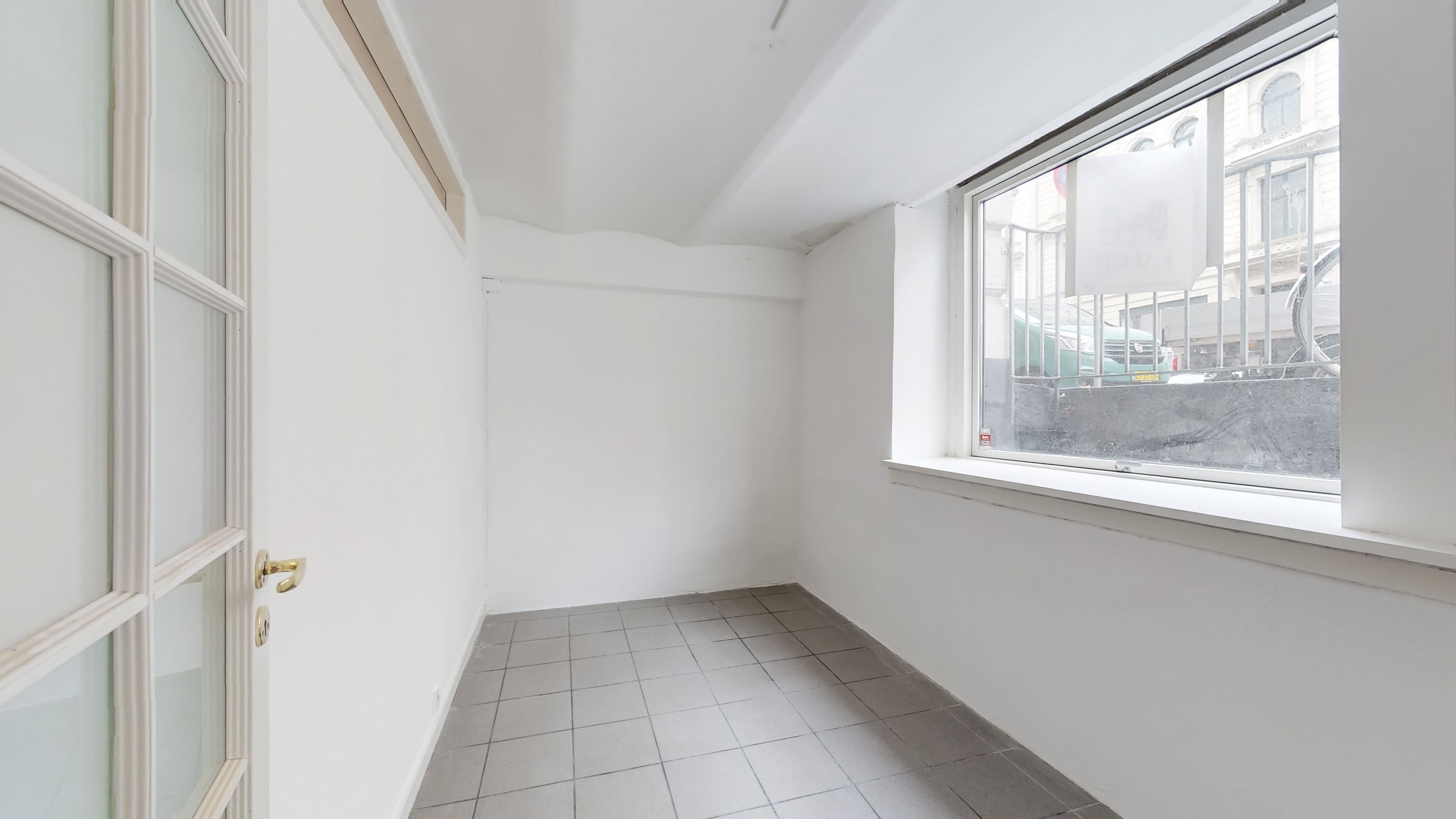 Et tomt rum med hvide vægge og gulv og en stor vinduesåbning med udsigt til en grøn bil og en høj bygning i baggrunden