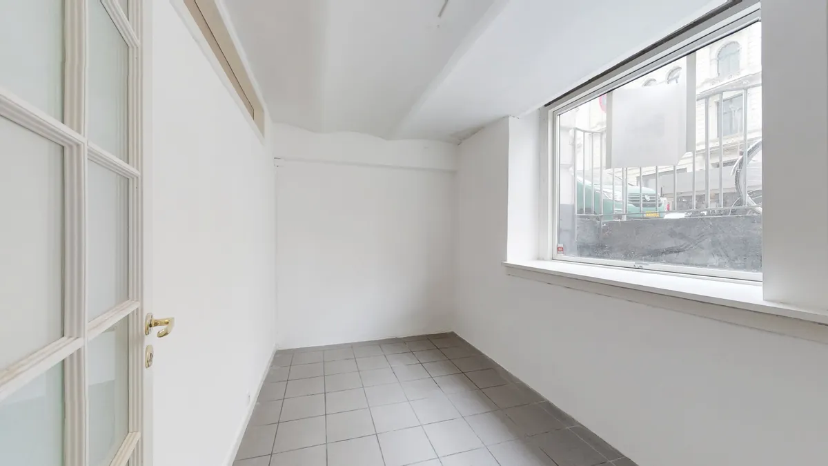 Et tomt rum med hvide vægge og gulv og en stor vinduesåbning med udsigt til en grøn bil og en høj bygning i baggrunden