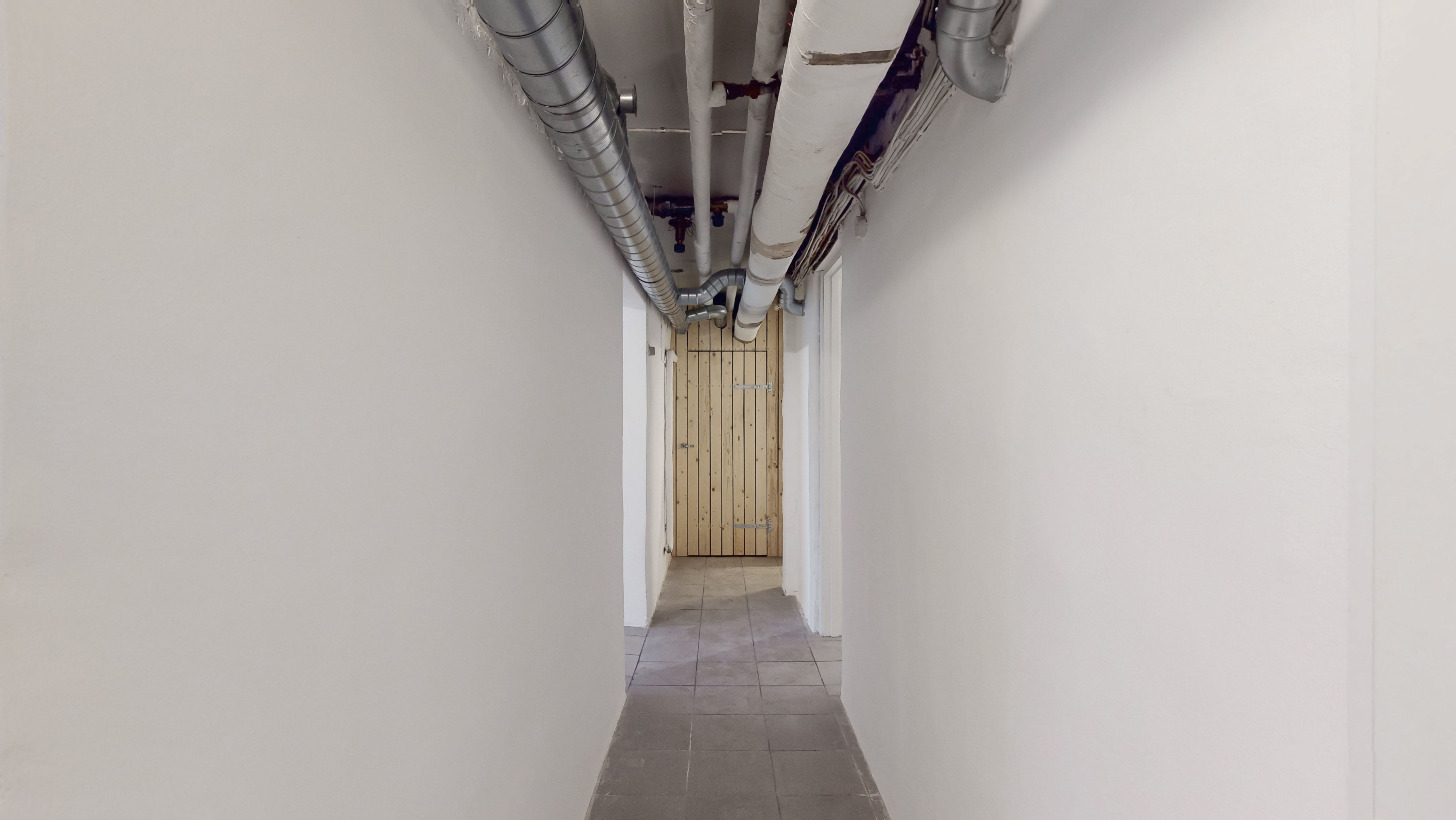 En række røretråde og metalrør er fastgjort til hvide vægge i en smal gang. En trædør er åben i væggen og giver adgang til en anden gang.