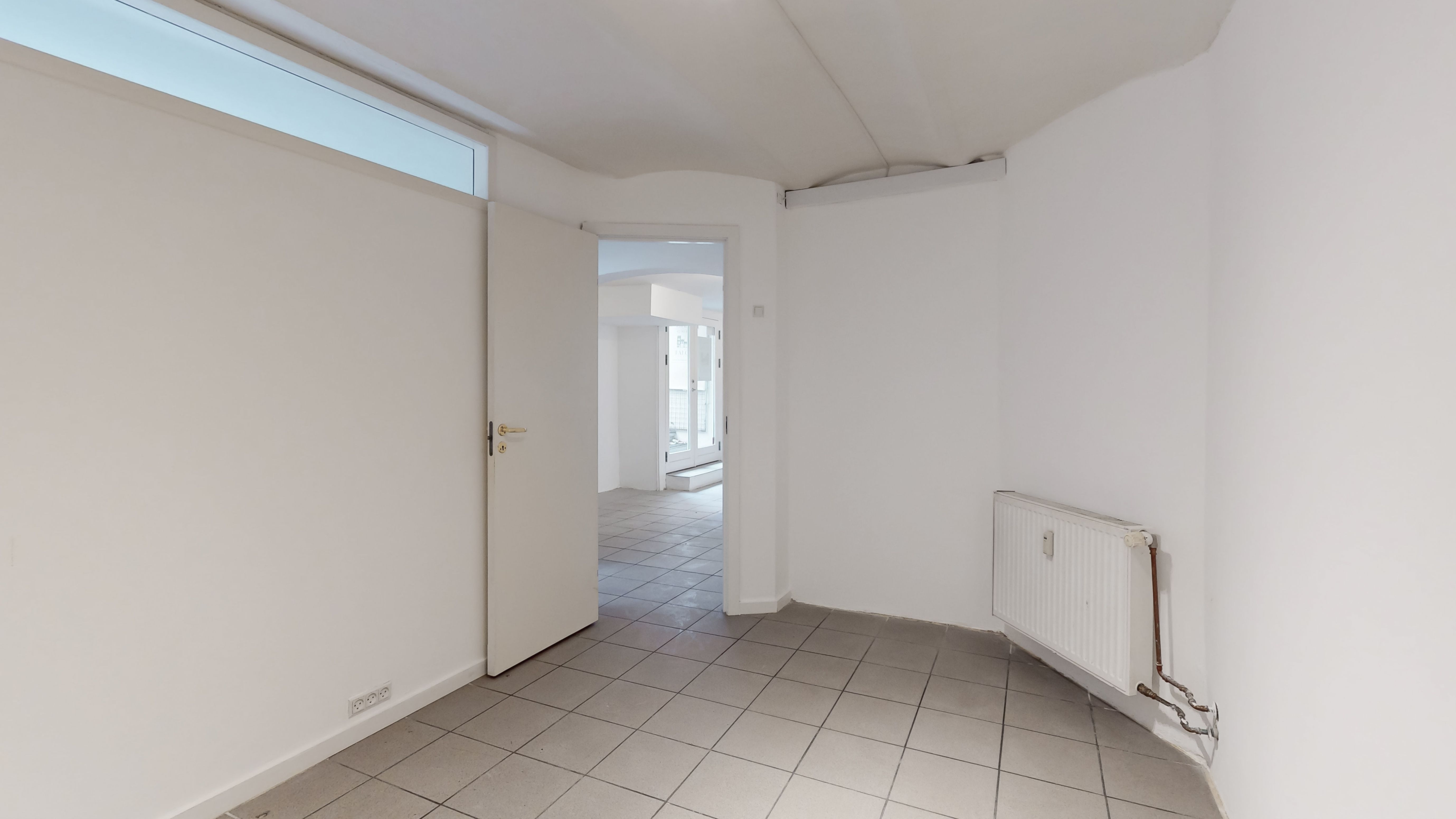 En tom og hvidmalet butikslokale med åben dør og tæt ved radiatoren og en indgangsdør med vinduesglas.