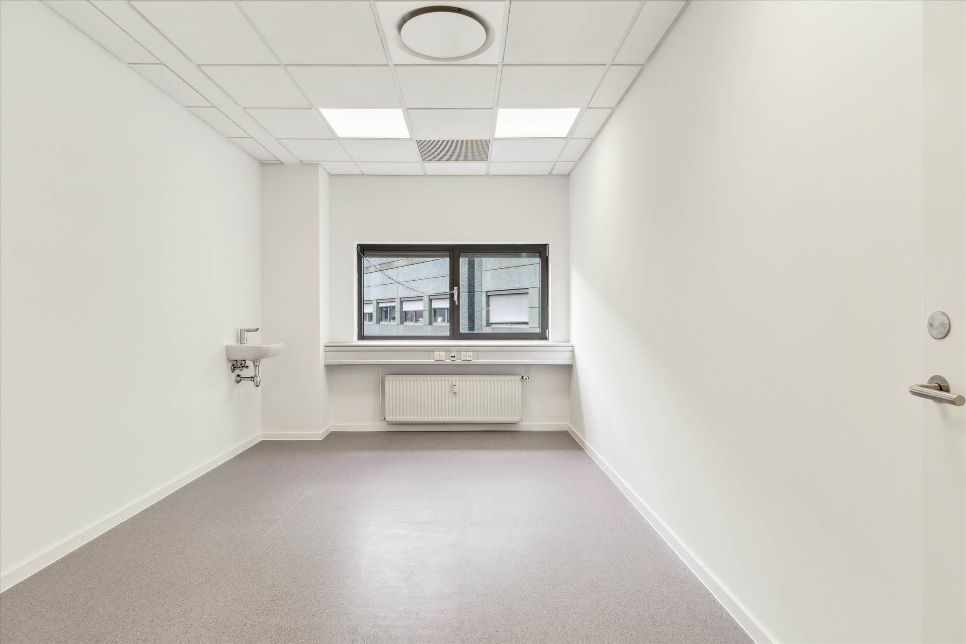 Lys, ren rum med hvide vægge og gulv og et vindue med skur. Et radiator er monteret på væggen under vinduet. Et håndvask med
