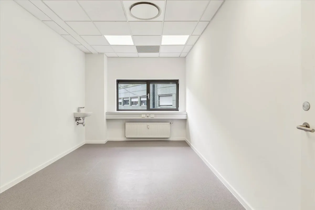 Lys, ren rum med hvide vægge og gulv og et vindue med skur. Et radiator er monteret på væggen under vinduet. Et håndvask med