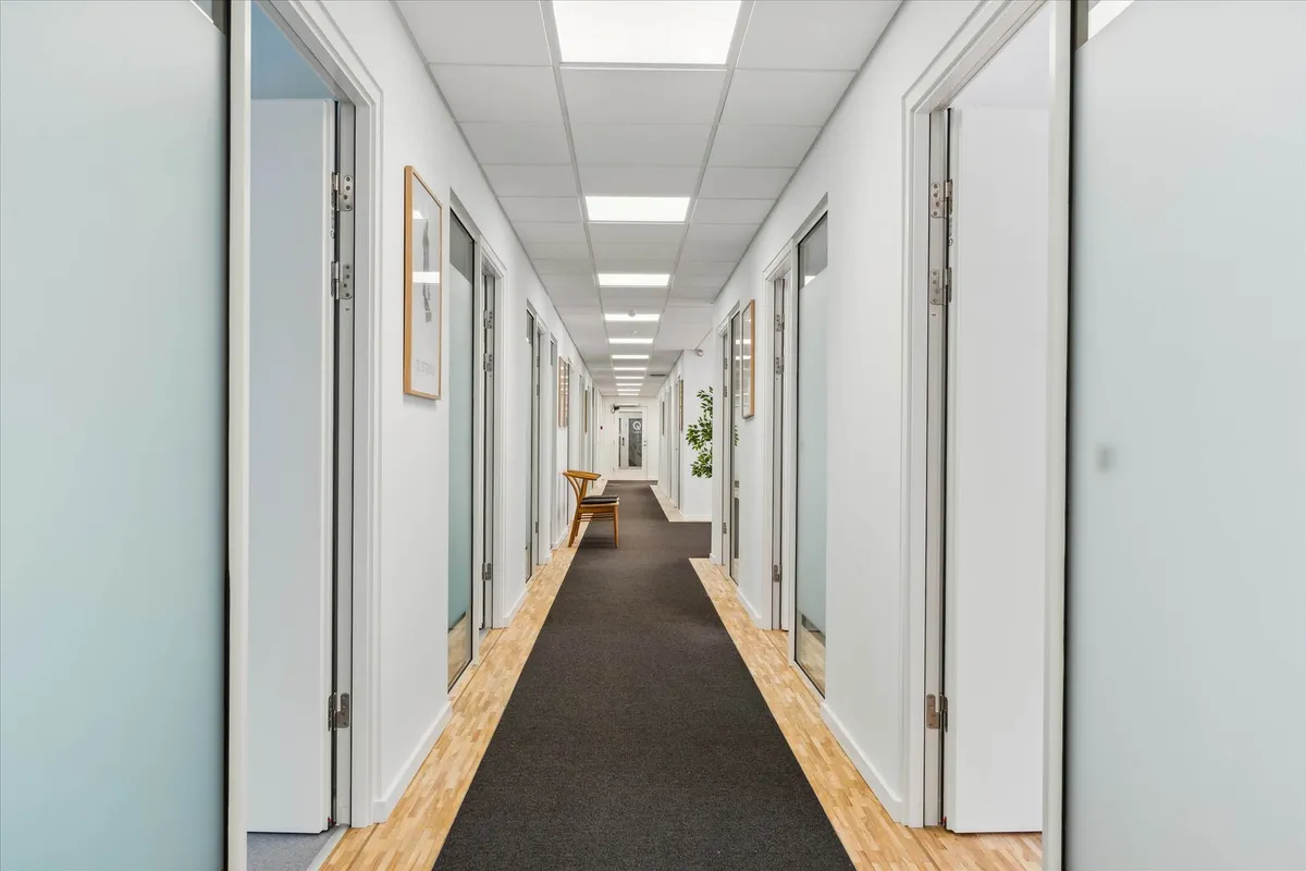 Langt, smal korridor med hvide vægge, trægulve og kunstig belysning i loftet. To døre med hjørneglas og møbler i en af dem