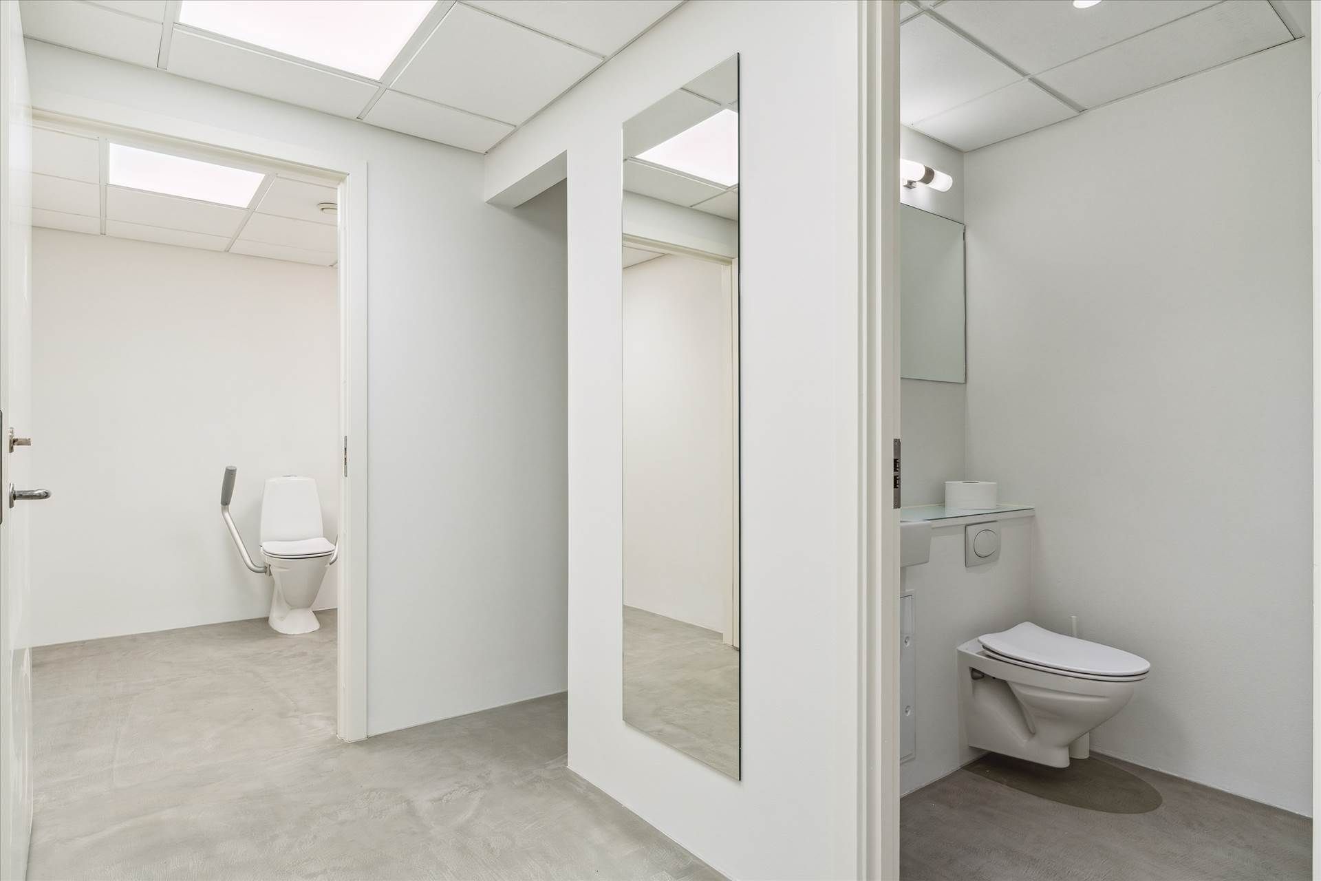 En hvid toiletrum med et toilet med håndtag og en spejl. Et toiletpapir står på et lille hylde og der er en lysekron over