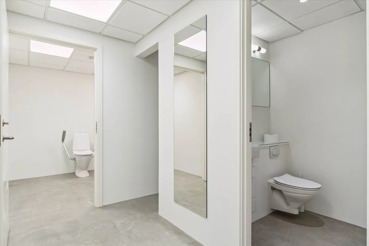 En hvid toiletrum med et toilet med håndtag og en spejl. Et toiletpapir står på et lille hylde og der er en lysekron over