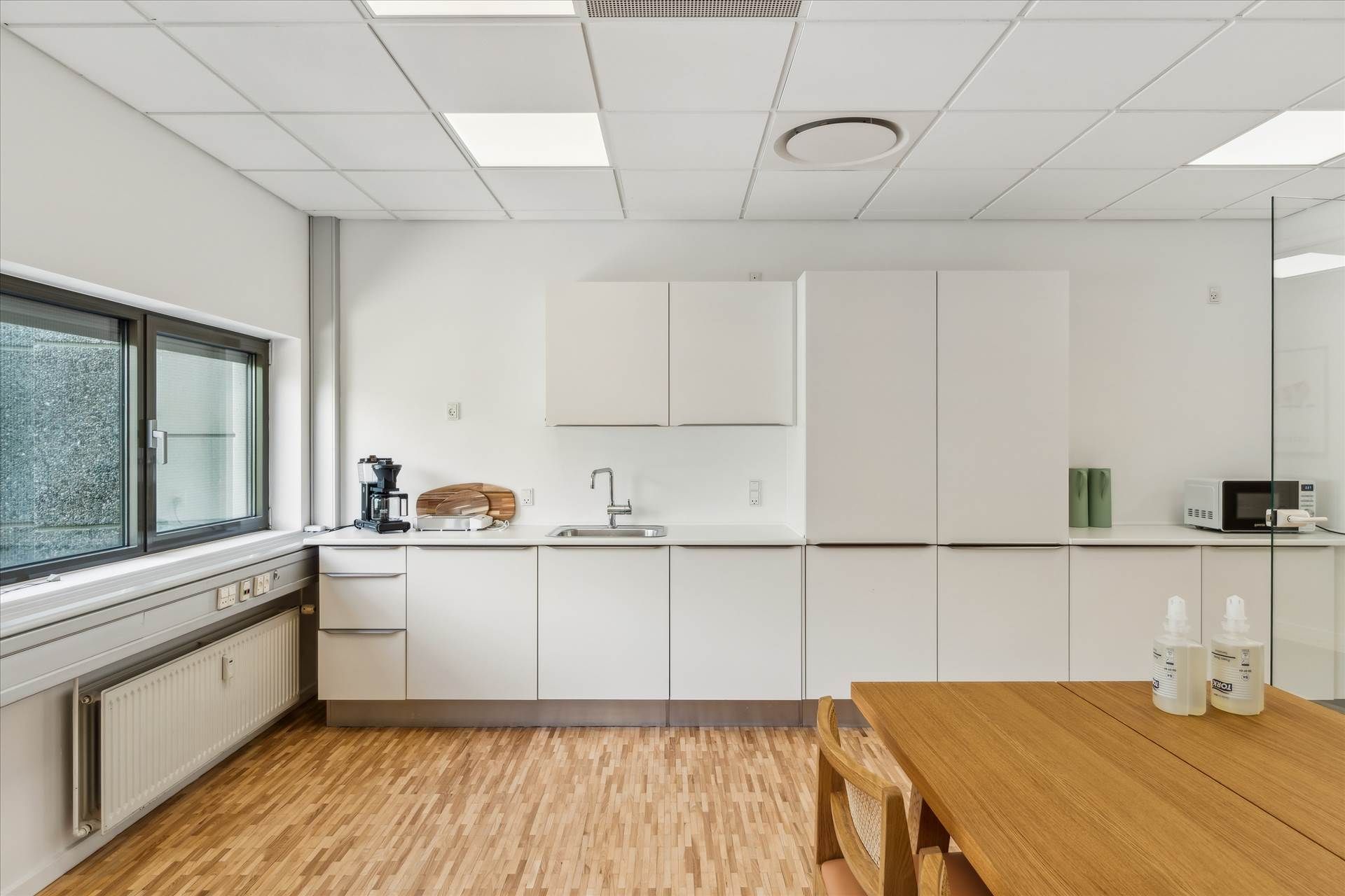 En moderne køkken med hvide vægge og trægulv, hvor der er en mikrobølgeovn og en kaffemaskine på en høj hylde.