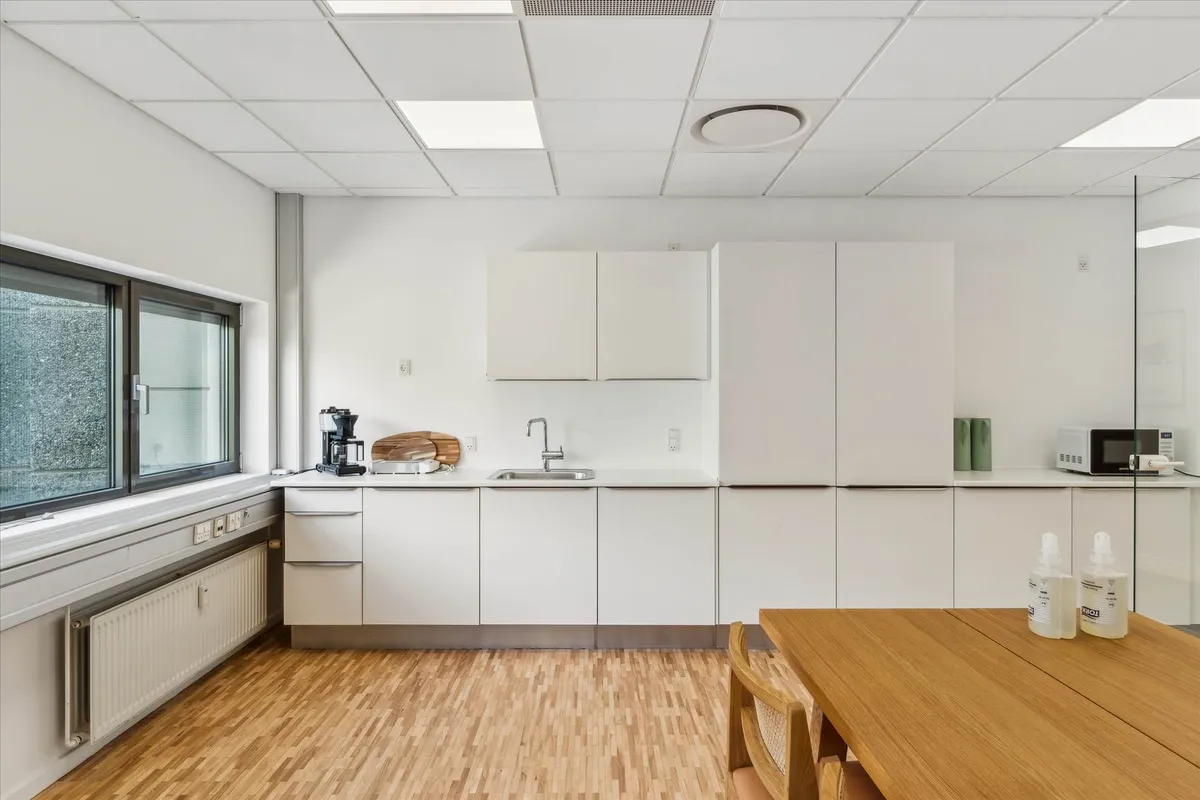 En moderne køkken med hvide vægge og trægulv, hvor der er en mikrobølgeovn og en kaffemaskine på en høj hylde.