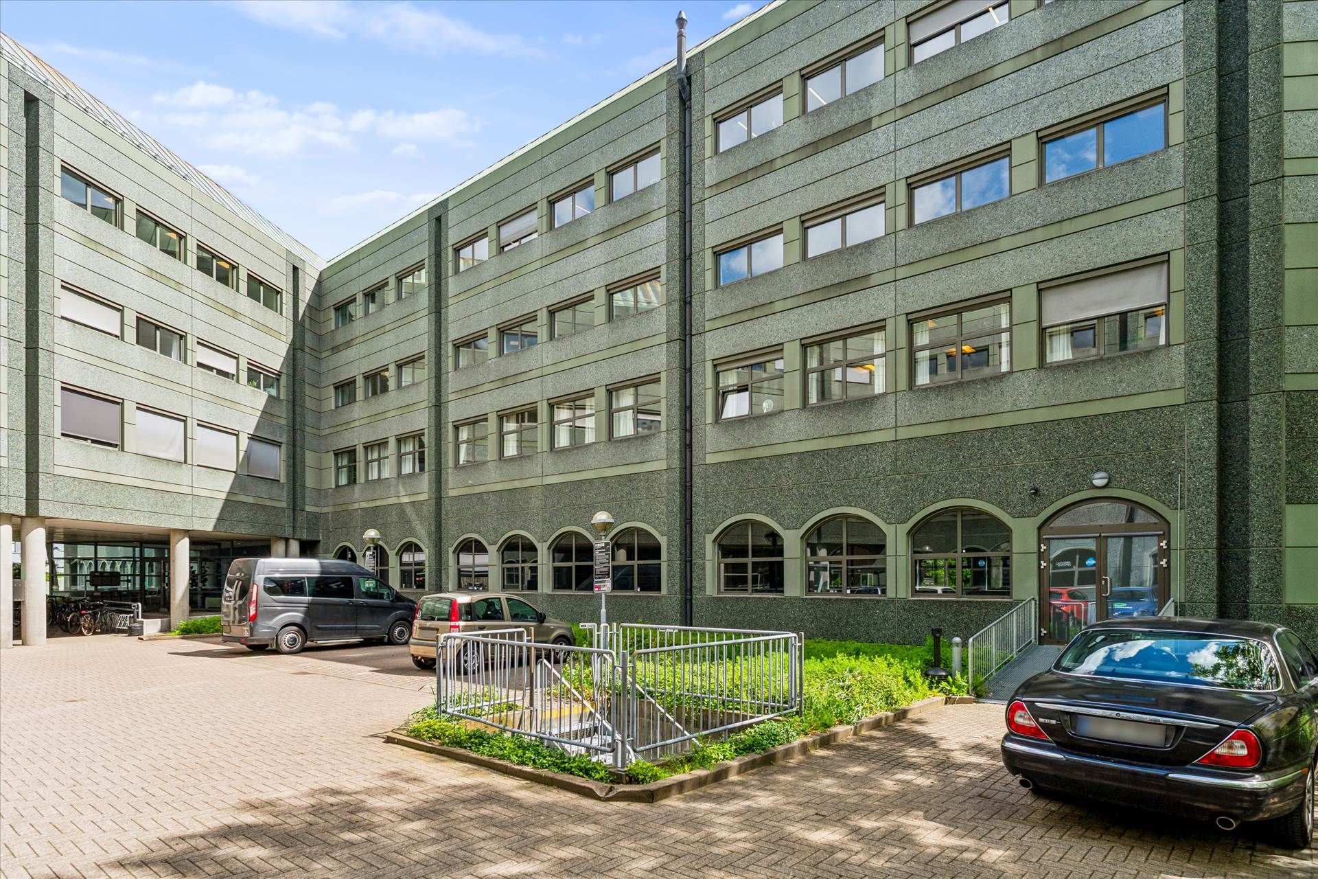 Det er en bygning med fem etager og mange vinduer, hvor der er parkeringsplads for biler.