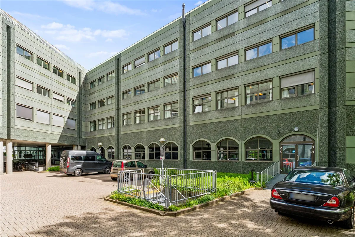 Det er en bygning med fem etager og mange vinduer, hvor der er parkeringsplads for biler.