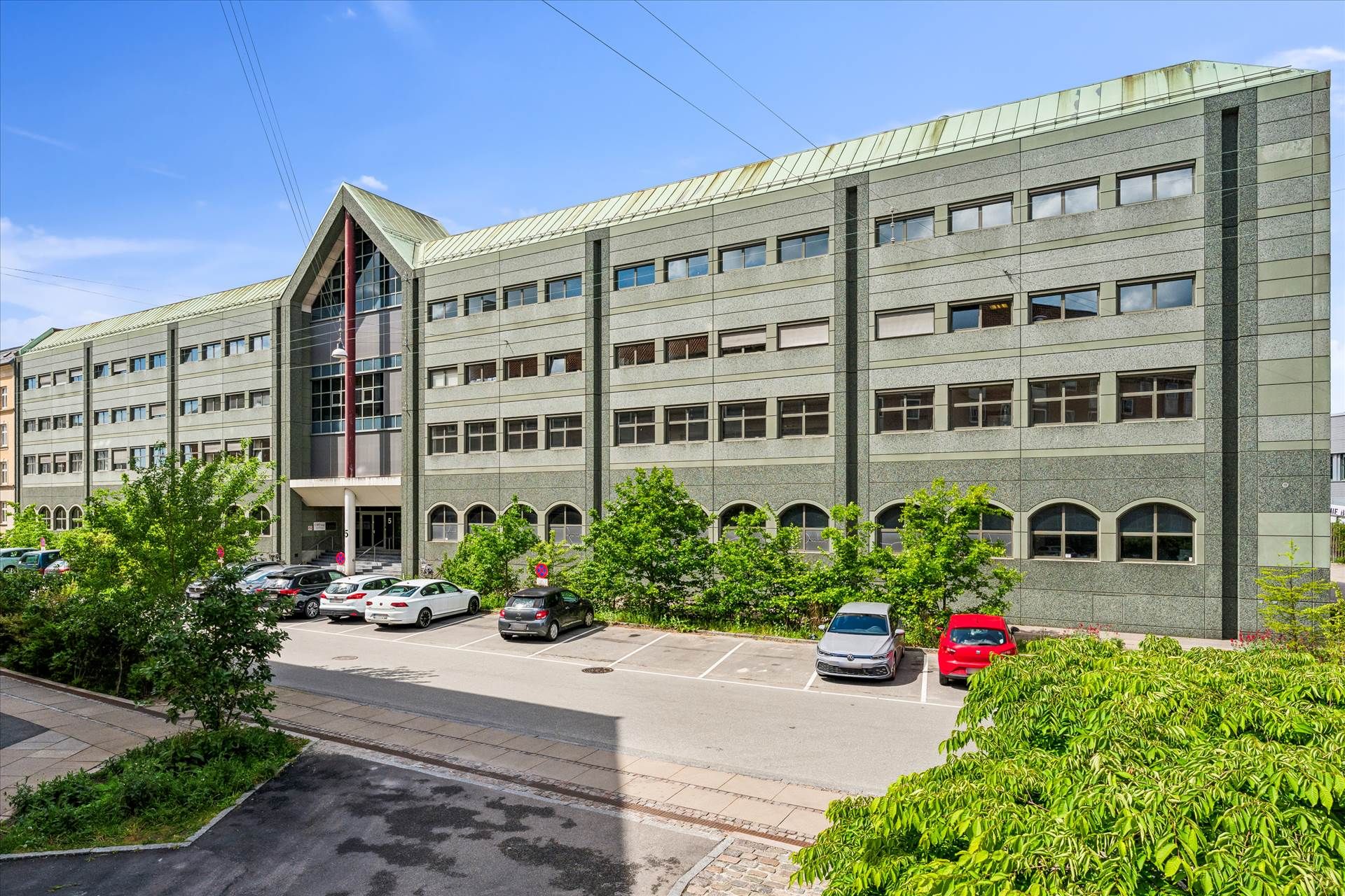 Bygning med flere etager, flere biler parkeret foran, lyseblå himmel, mange vinduer, grønne planter og træer.