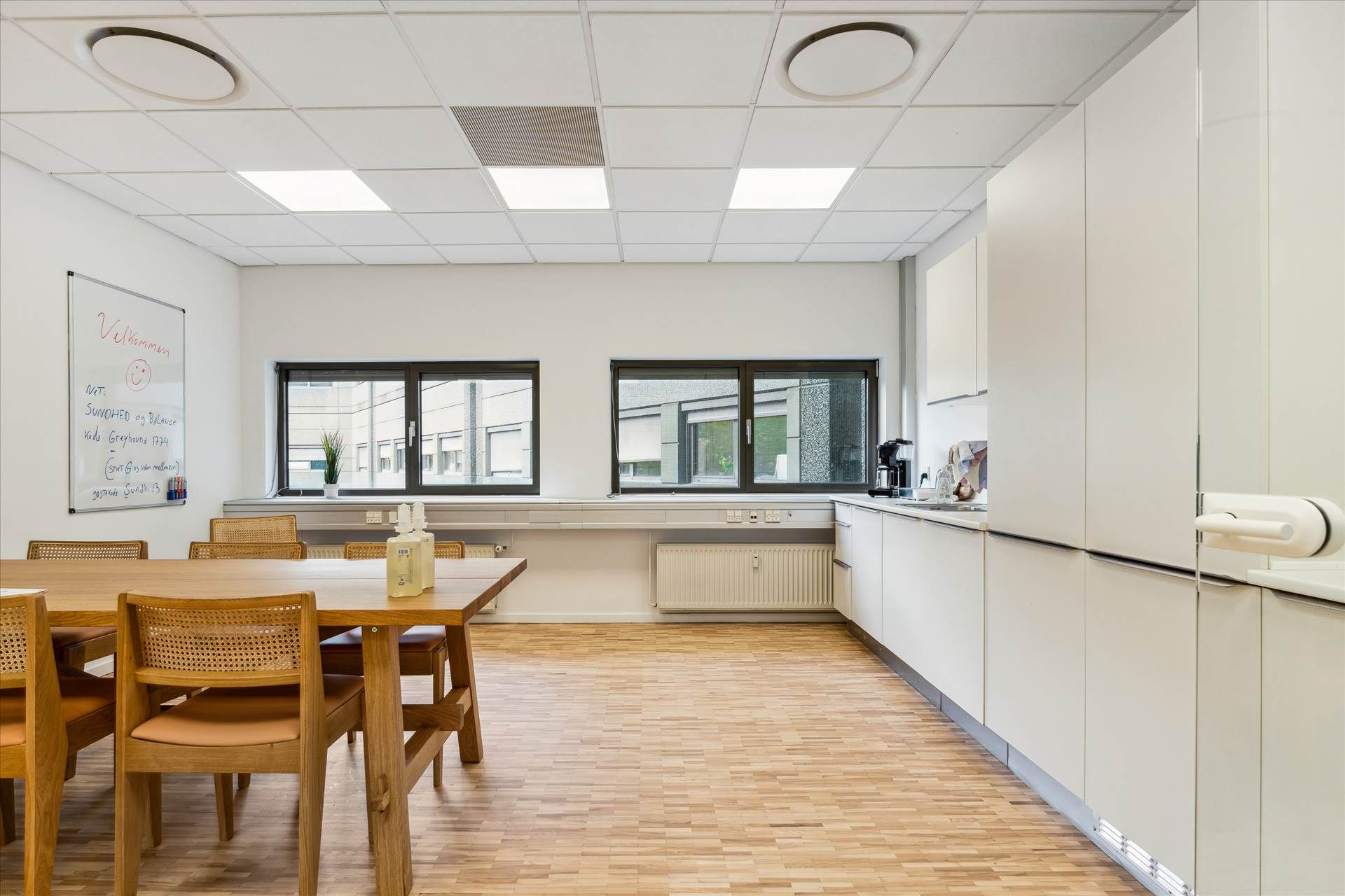 I et moderne mødelokale er der en stor bord, hvor der er sat flere stole rundt. Der er en hvid væg med en tavle og et