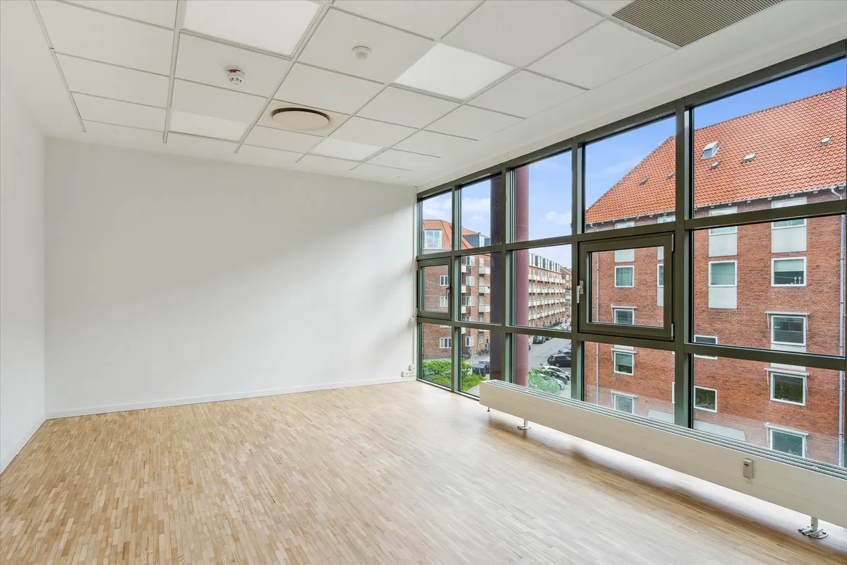 Tomt kontor med lyse trægulve, hvide vægge og store vinduer med udsigt til gaden og bygningerne