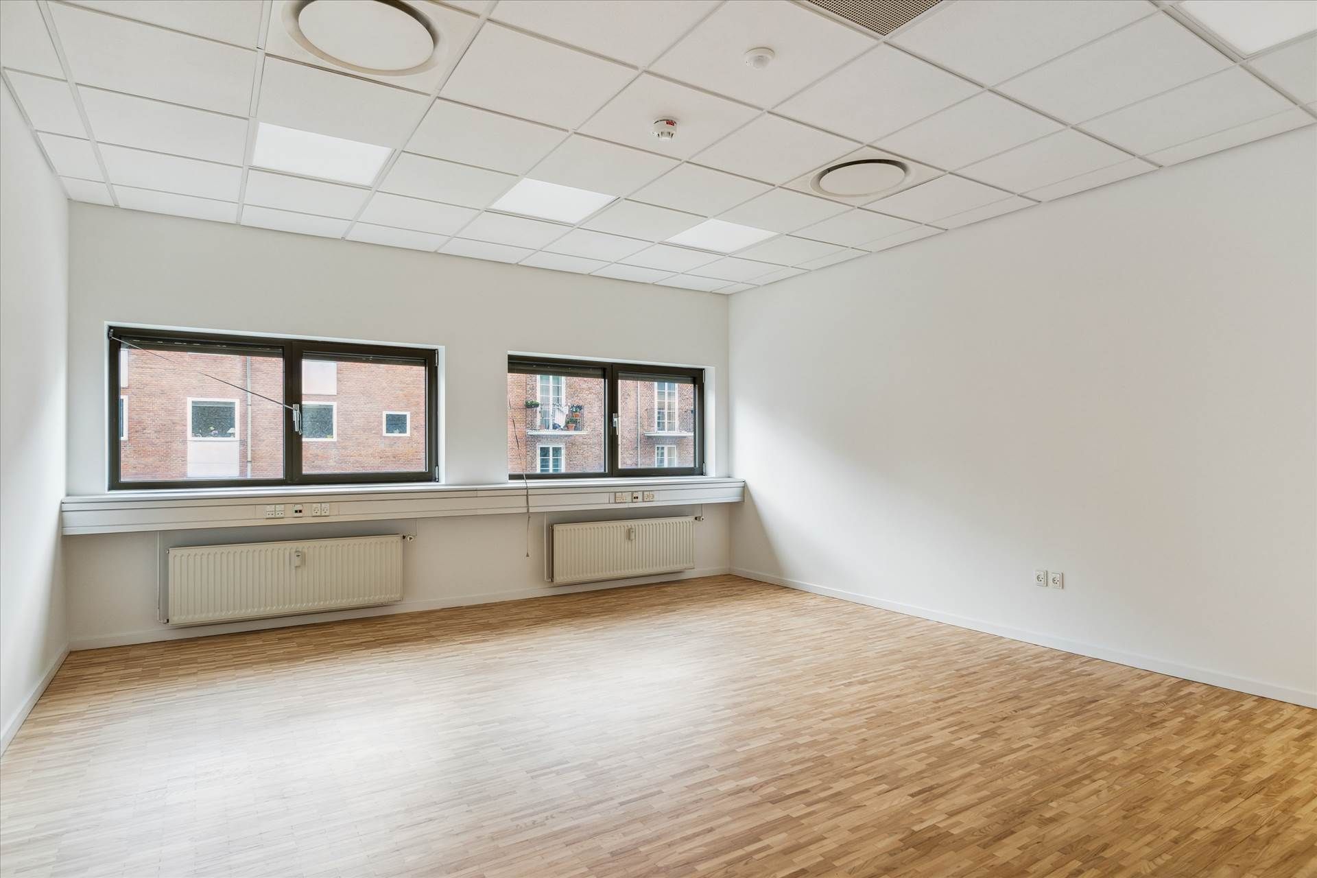 En tom mødelokale med hvide vægge, trægulv, store vinduer og radiatorer under vinduet. Gulvet og loftet er lyst og rummet