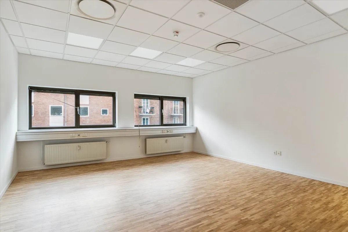 En tom mødelokale med hvide vægge, trægulv, store vinduer og radiatorer under vinduet. Gulvet og loftet er lyst og rummet