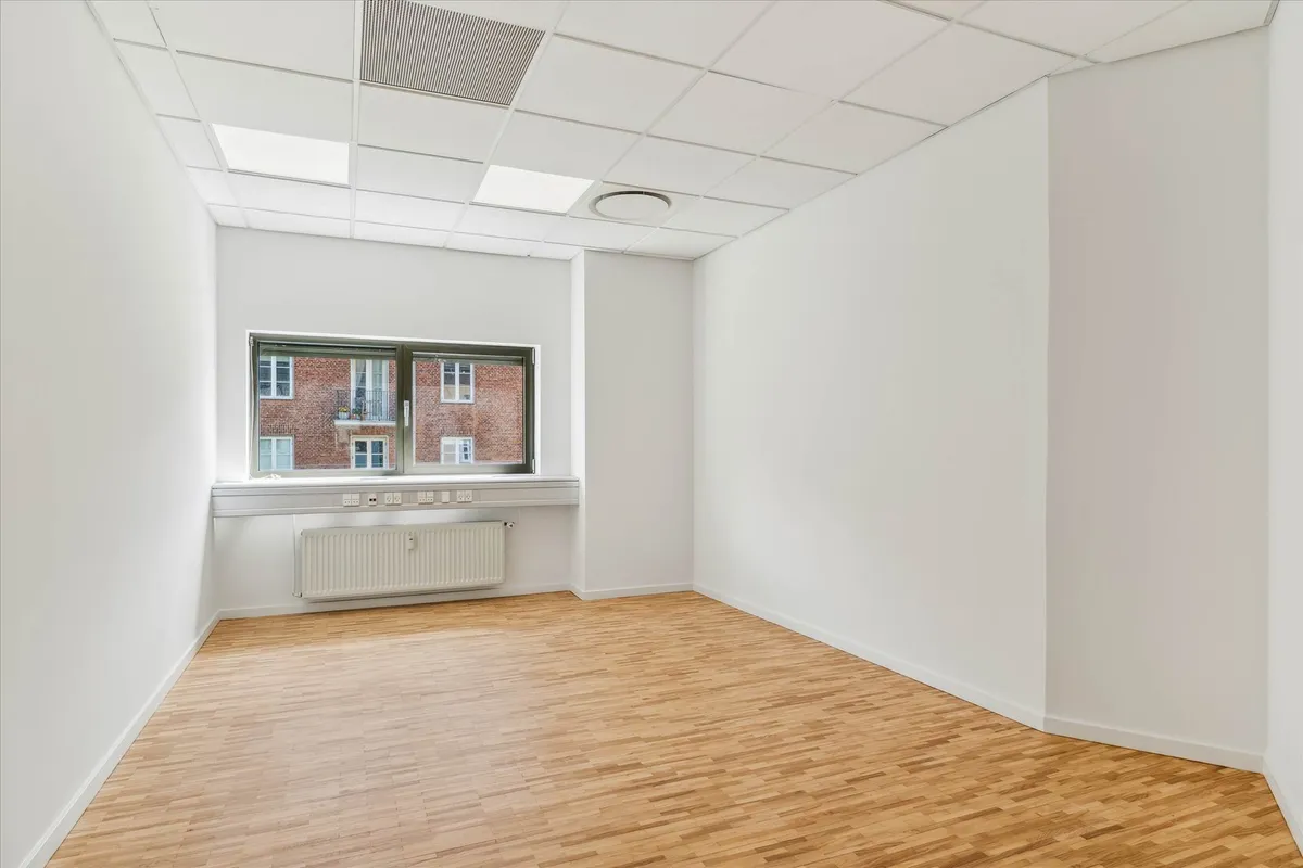 En tom kontorværelse med hvide vægge og gulv samt store vinduer og et hvidt radiatorpanel langs væggen