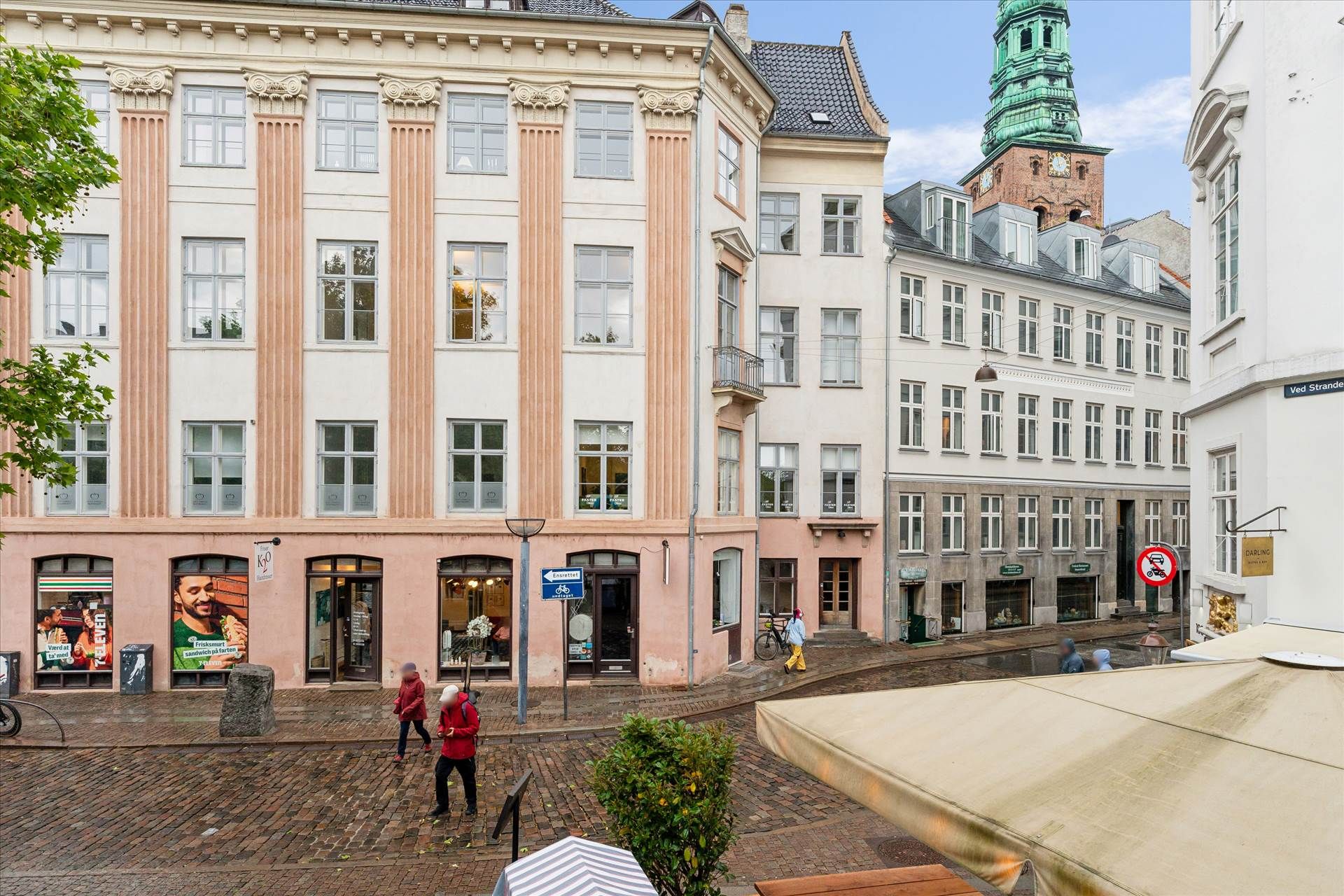 En smal gade med små butikker og mennesker, der går, samt en søjle med en skilt og et bord med en paraply.