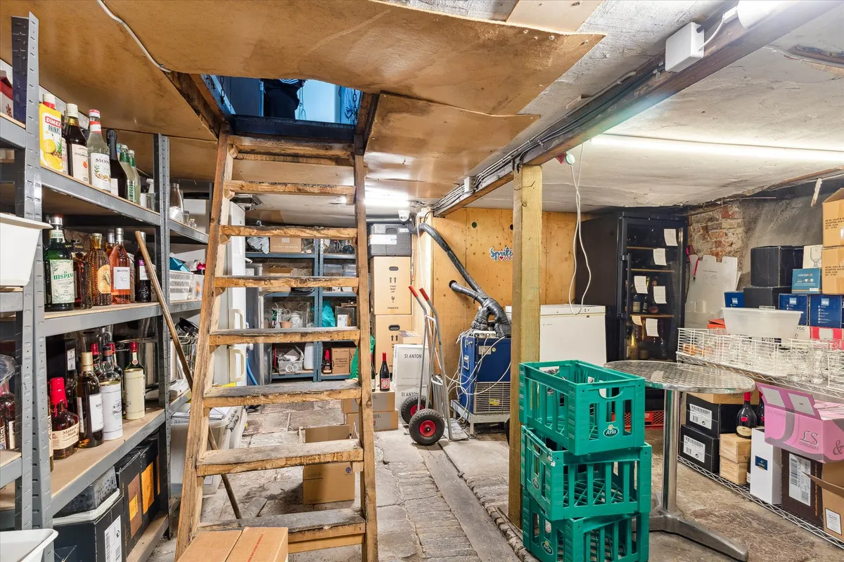 En vinkælder med lagerhylder, flasker og etiketter, flere kasser og kassevogn samt to borde med en række vinflasker.