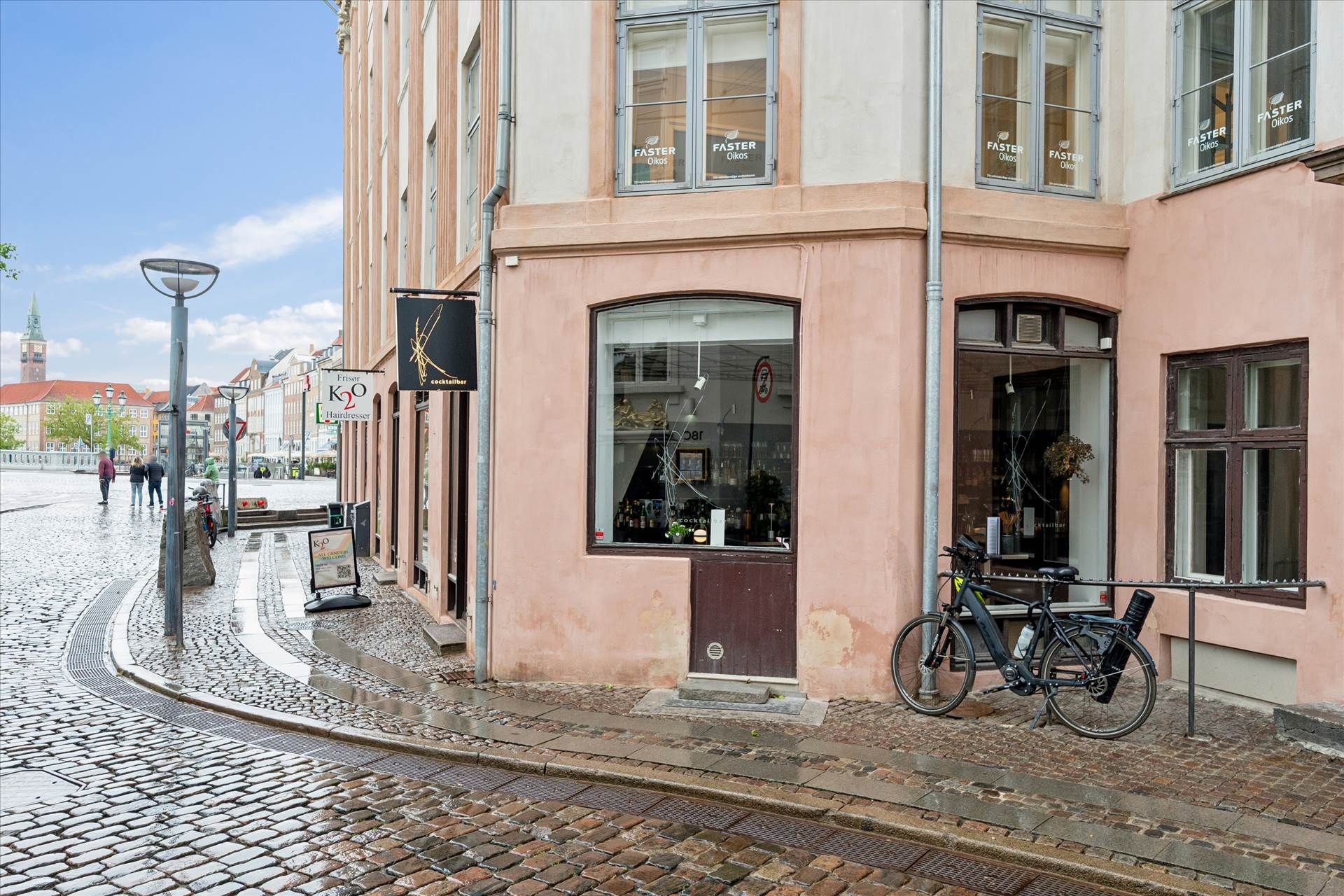 En cykel parkeret foran en K20 frisør. Der er flere mennesker på gågaden. Bygningen har flere vinduer og en skilt med Faster