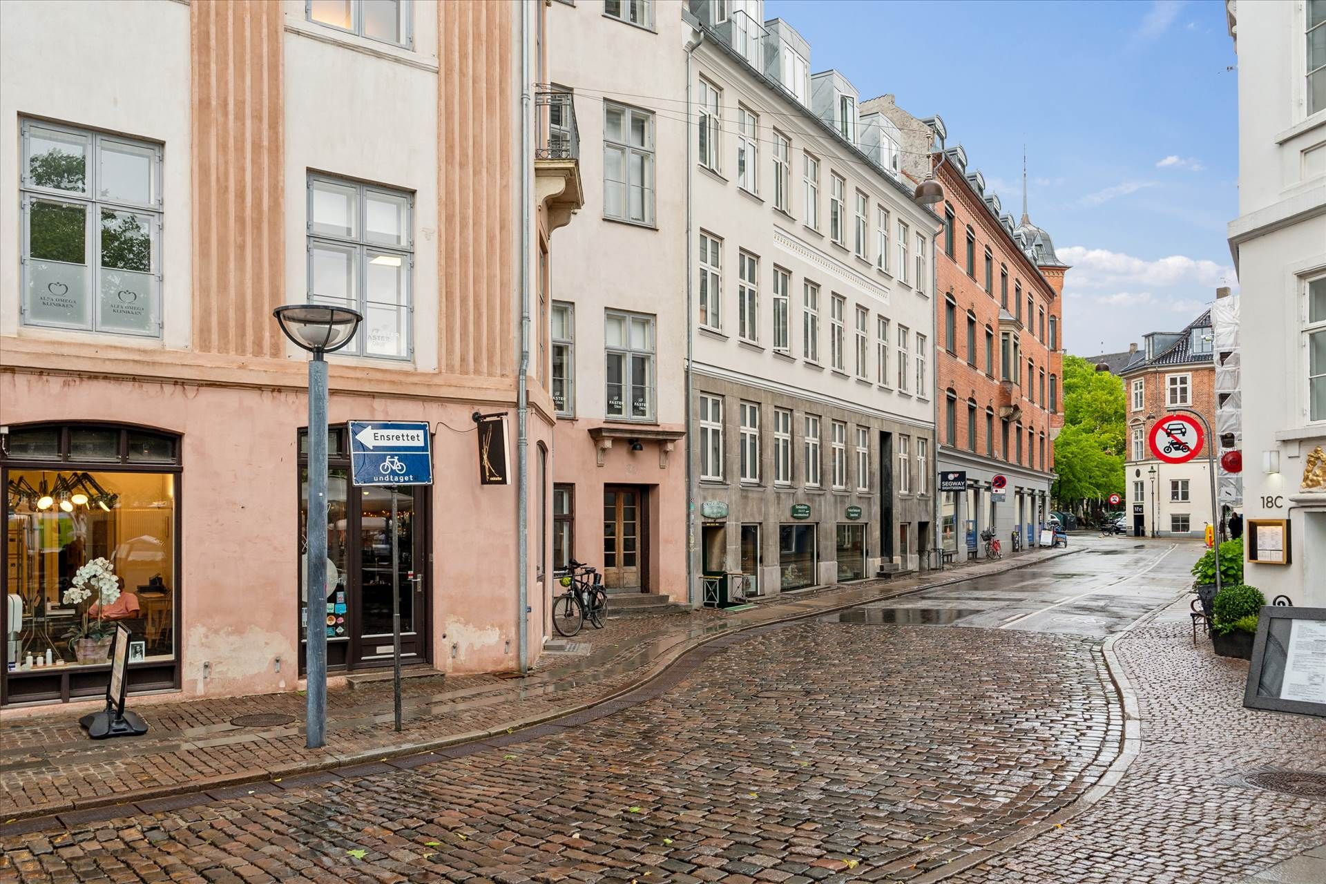 En tom gade med stenbrolægning og et lille skrånende område med en cykelparkering, der viser en grøn skilt med et forbudstegn