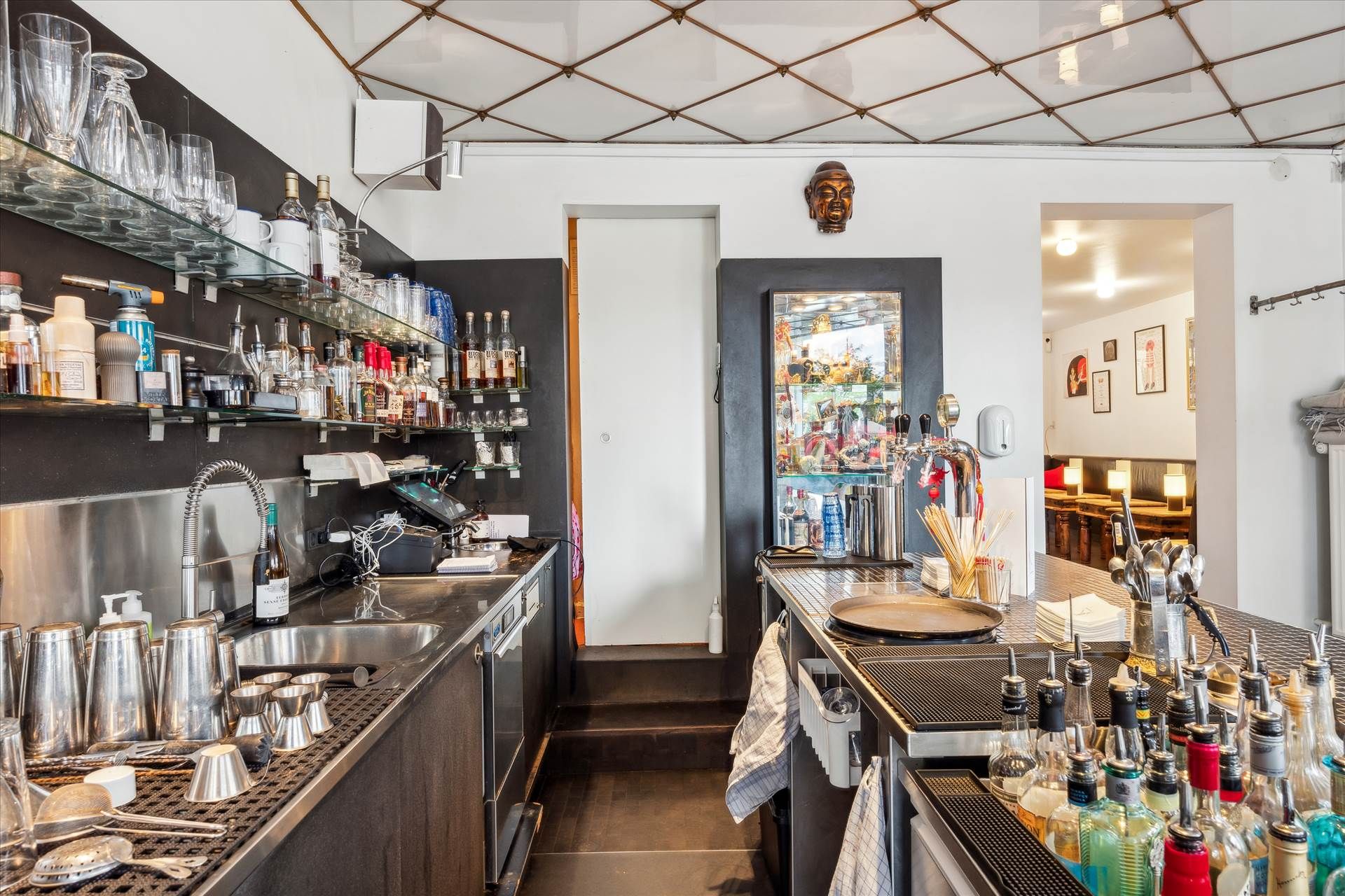 Glas og flasker på hylder, en åben dør, et spejl, et glasvæg og en maskine på en bar i et lokale