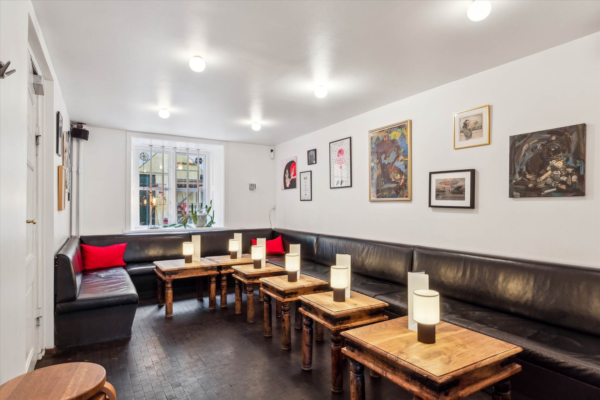 Indretning med to lædersofaer med røde puder og lysestager, udstilling af malerier på hvide vægge og gulvbelægning i træ