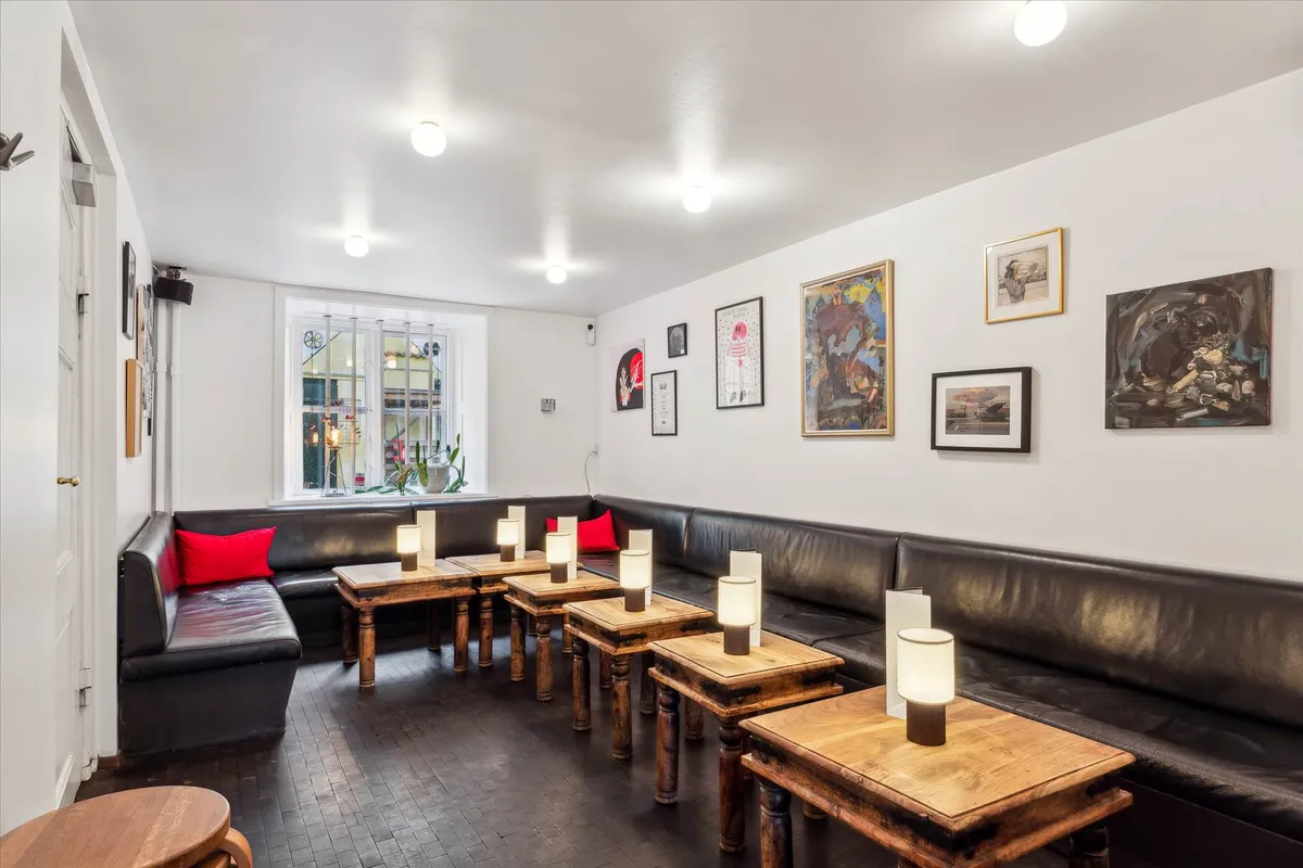 Indretning med to lædersofaer med røde puder og lysestager, udstilling af malerier på hvide vægge og gulvbelægning i træ