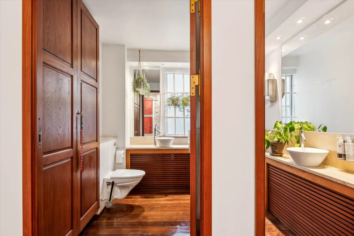 En moderne toiletrum med dobbelt vask og toilet, to hængeplanter og et lille vindue med udsigt til gaden