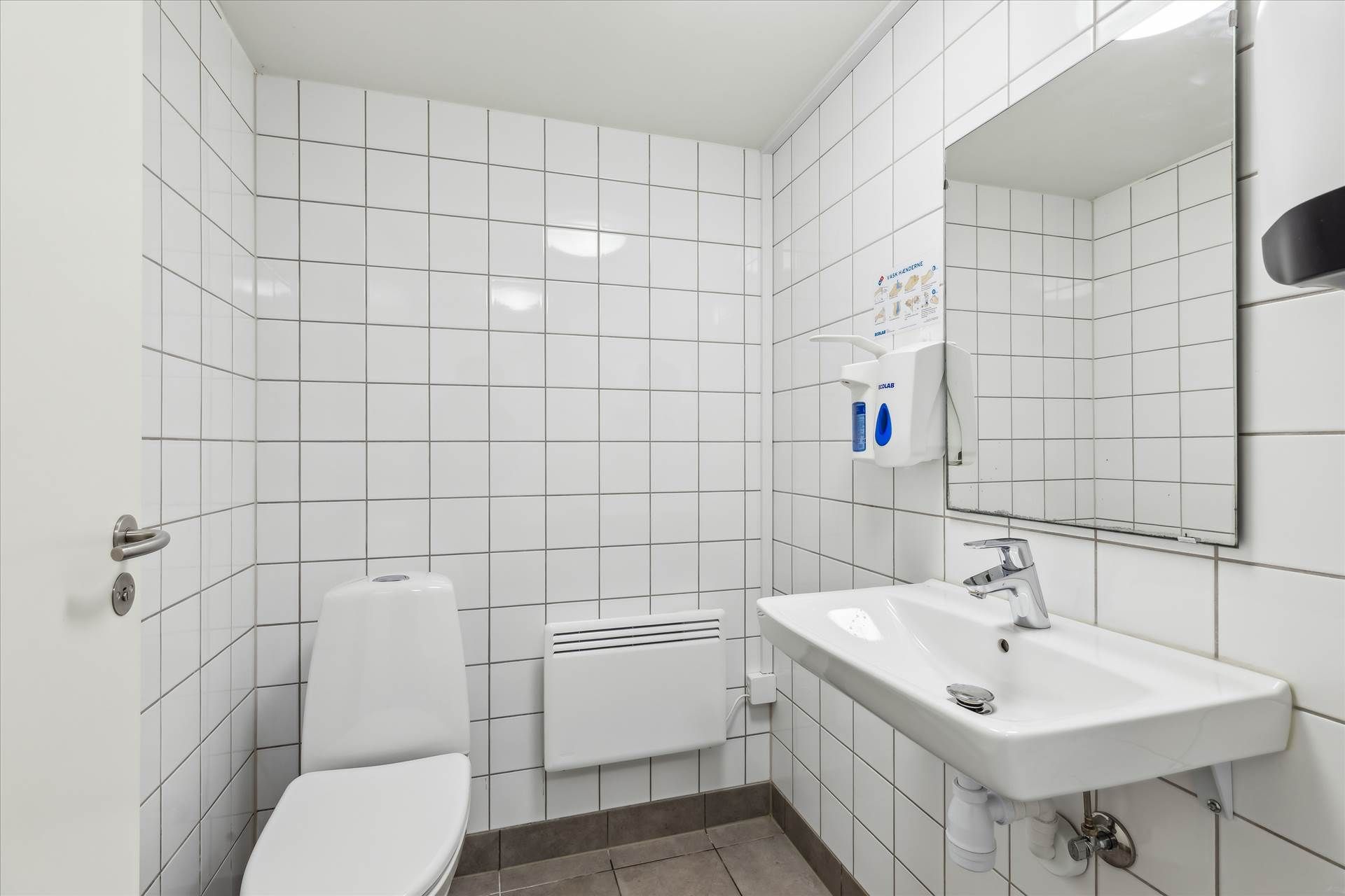 Rent og pænt badeværelse med toilet, hvid tøjvask, håndvask, spejl og hånddesinfektionsdispenser. Der er en hvid