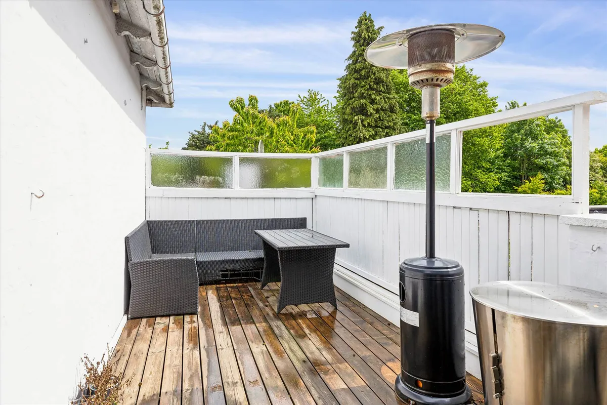 En terrasse med et brun sofa, et bord og en spisestue. Der er en have med træer og en varmelampe.