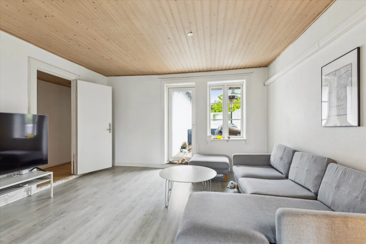 Grå sofa i stuen med rundt hvidt bord og tv-skærm med en udgang til et køkken