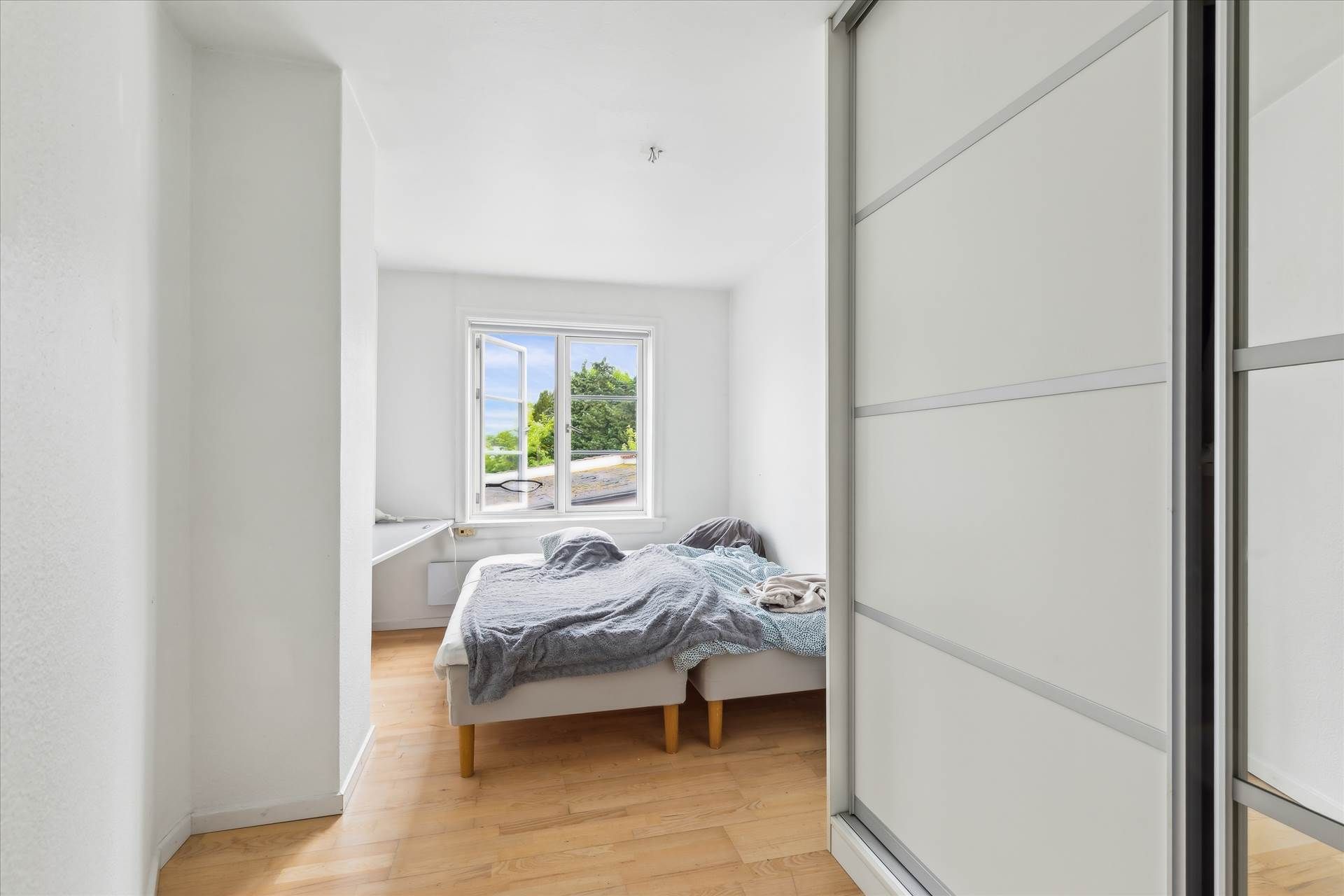 Et soveværelse med trægulv og hvide vægge, et enkelt dobbeltseng med tæppe og pude og en åben vindue med udsigt til en have.