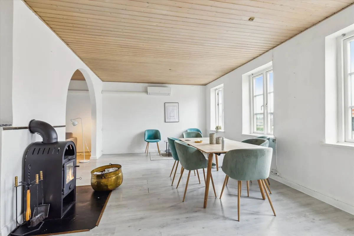 En indretning med hvide vægge og loft, et stort træbord med stole, en gulvlampe, et komfur, og vinduer med udsigt til byen