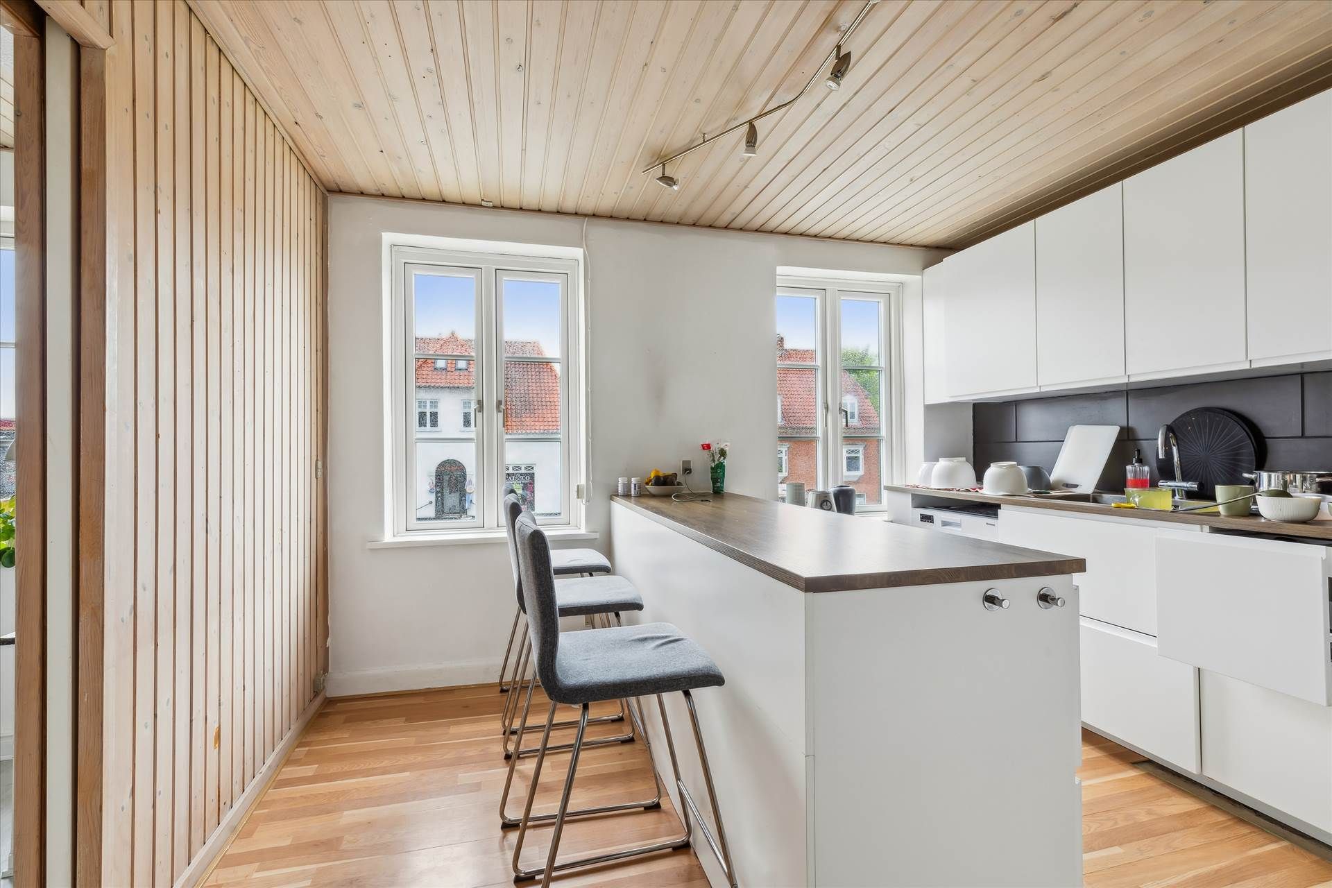 En moderne køkken med hvidt loft, lysegrå stole, en høj bar, og vindu med udsigt til en bygning med rødt tag