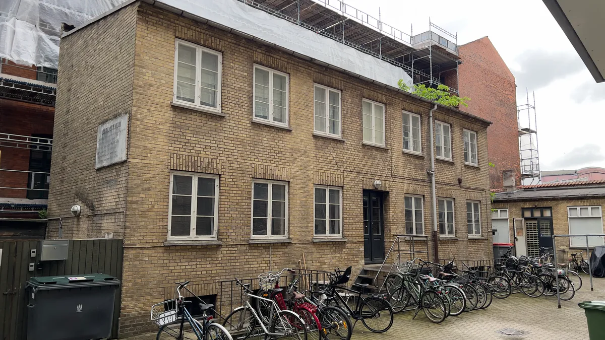 En række cykler er parkeret udenfor et lejet lokale i en by. I baggrunden er der bygninger med stige og skraldspande