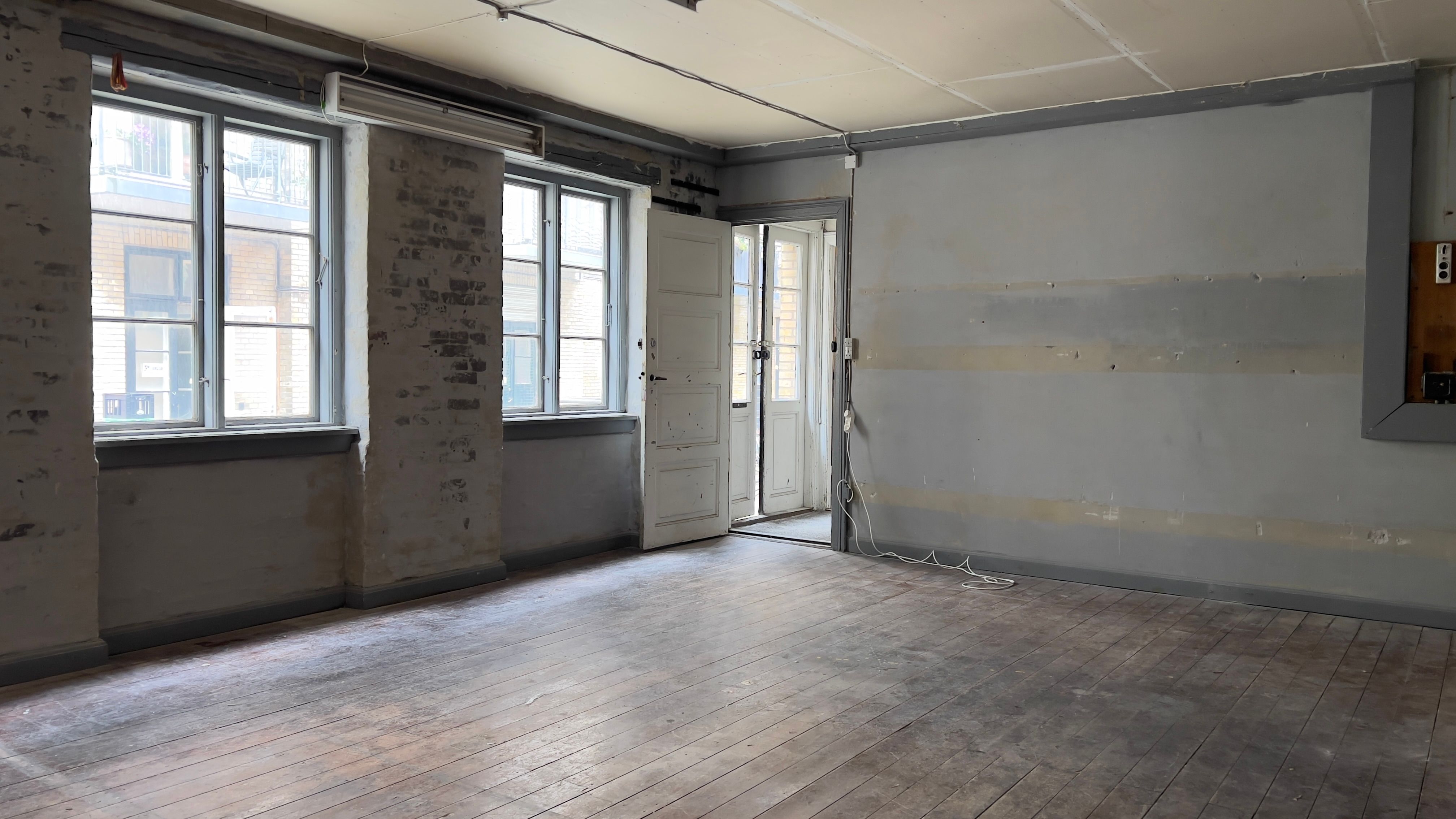 Tomt kontor med trægulv og åben dør til siden og vinduerne med udsigt til en bygning og balkoner