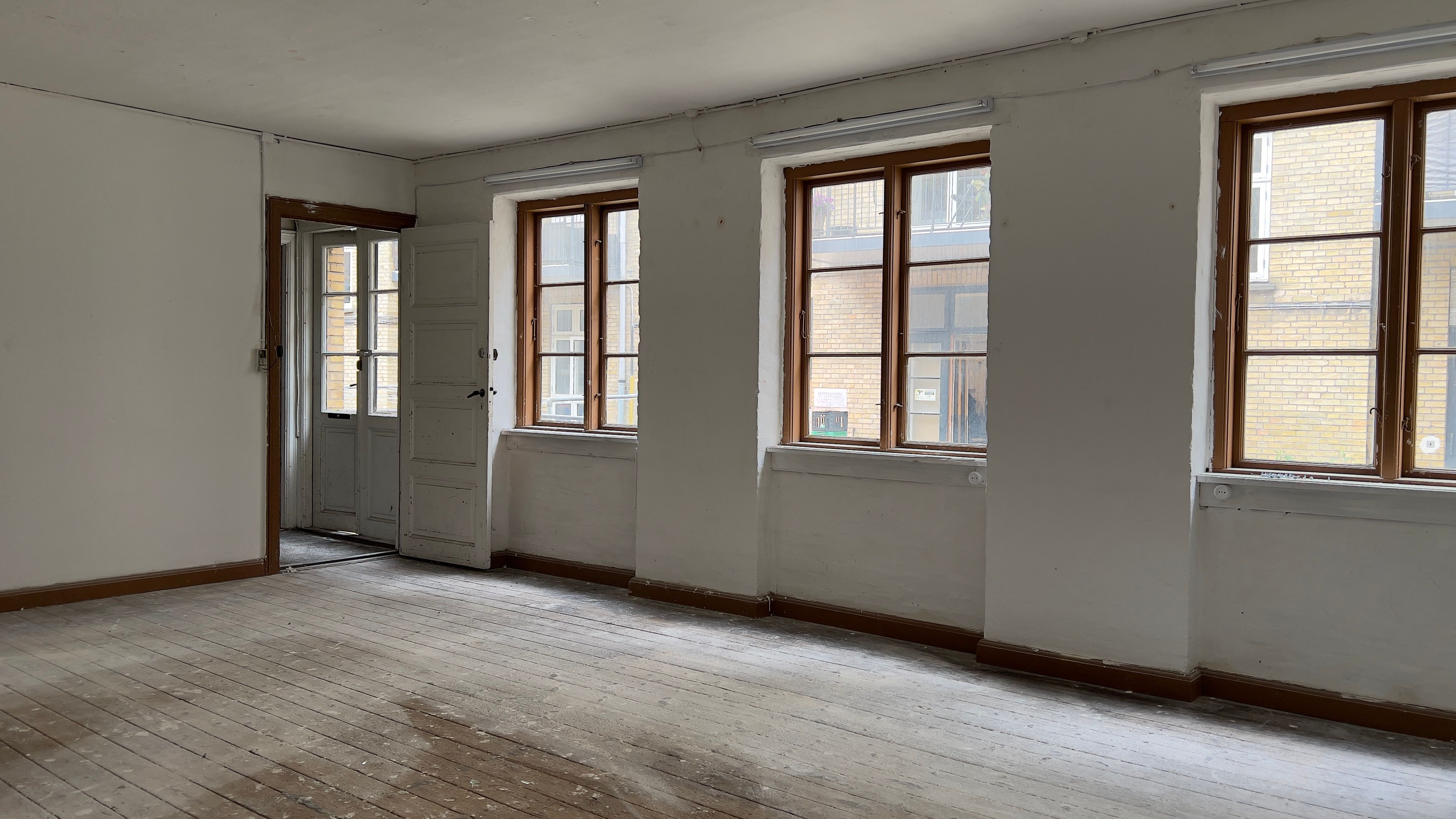 En tom kontorrum med trægulv og lyseruder mod gaden og tre store vinduer med højt vindueskarm