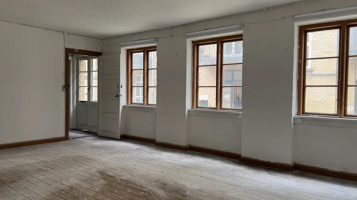 En tom kontorrum med trægulv og lyseruder mod gaden og tre store vinduer med højt vindueskarm