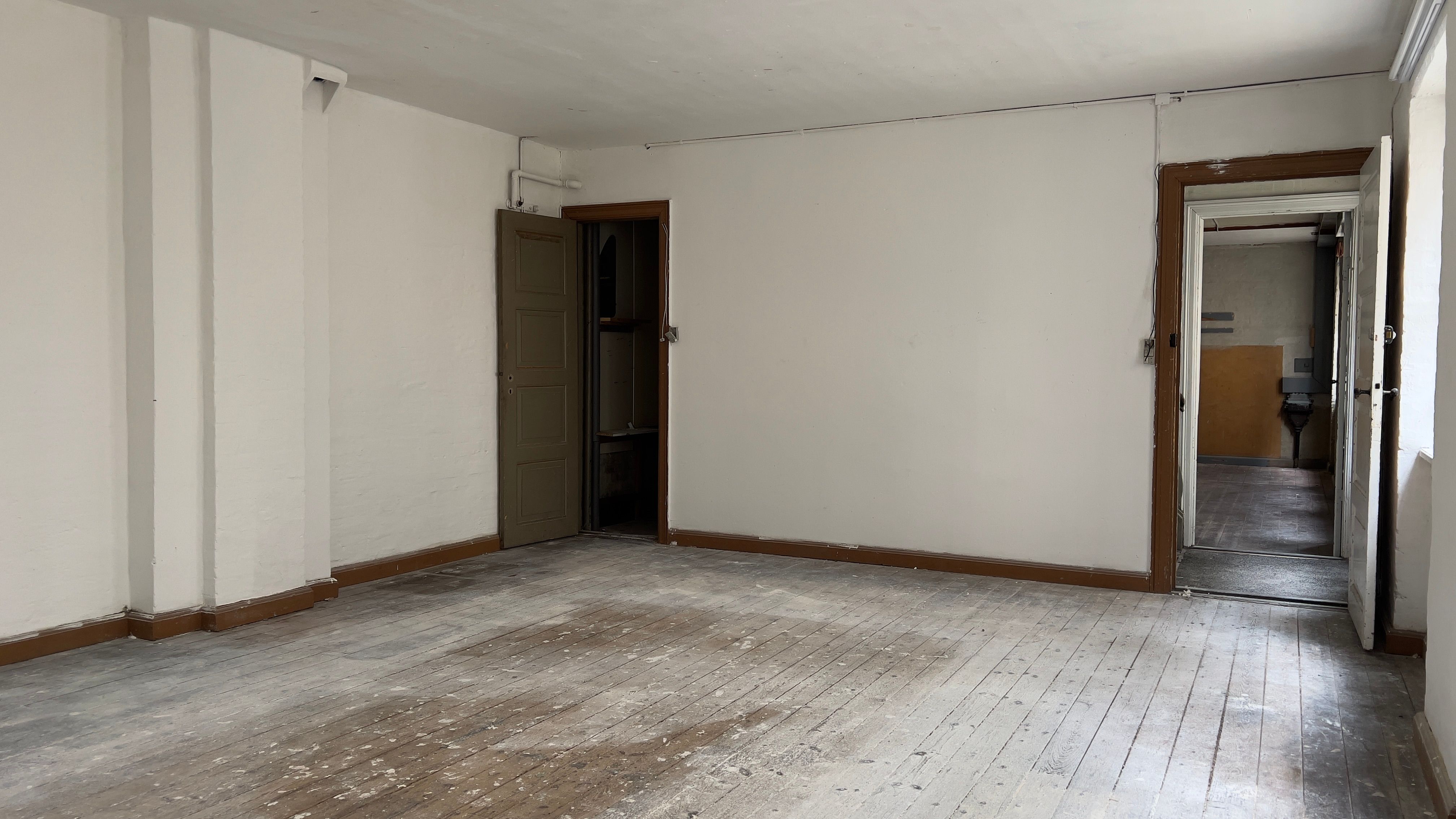 En tom og ren stue med åbne døre til siden og gulvet og væggene er træfarvede og har nye hvide malingsspor.