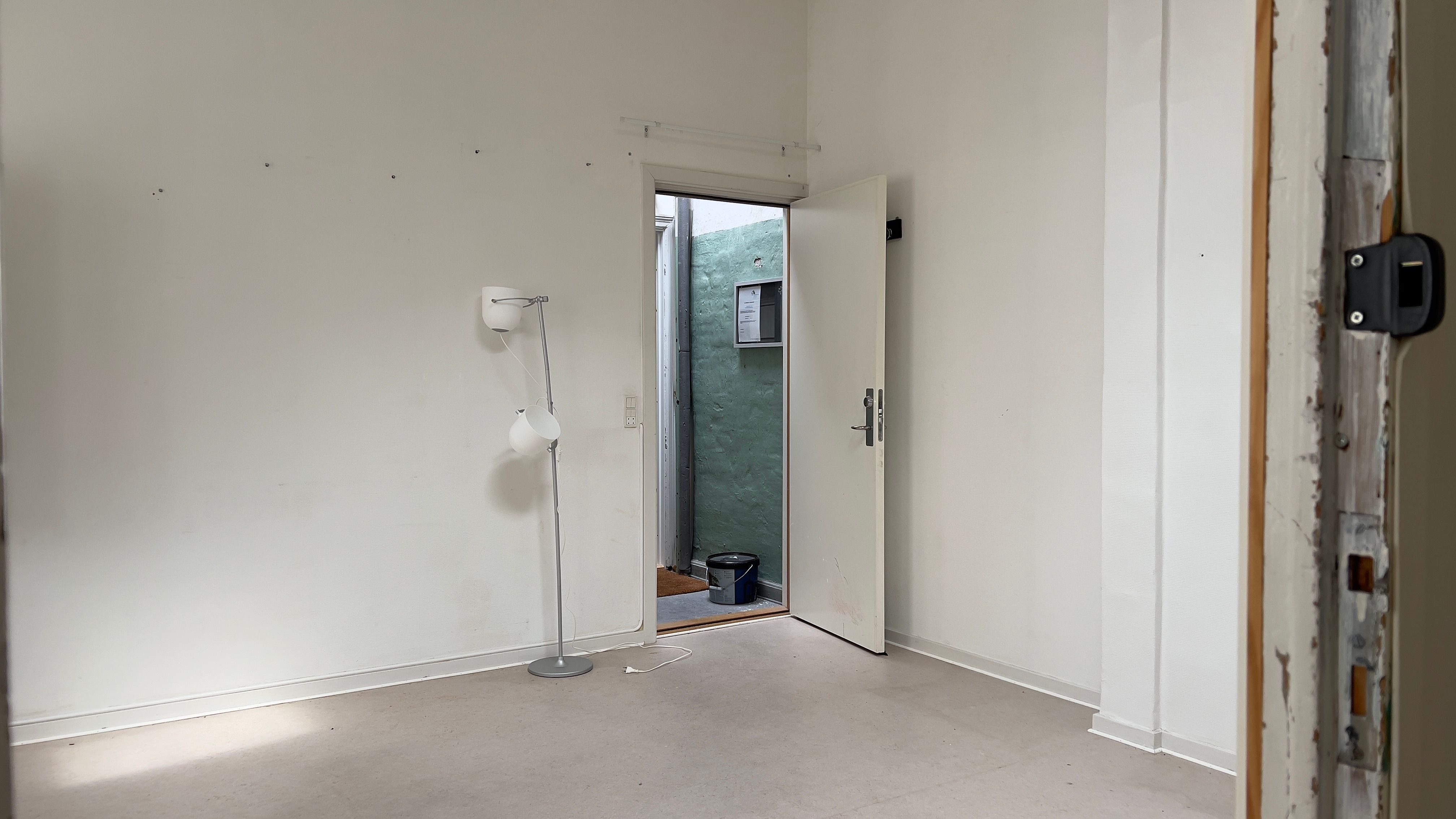 En tom stue med en hvid væg og et åbent hvidt dørblad, en lampe og en bøtte