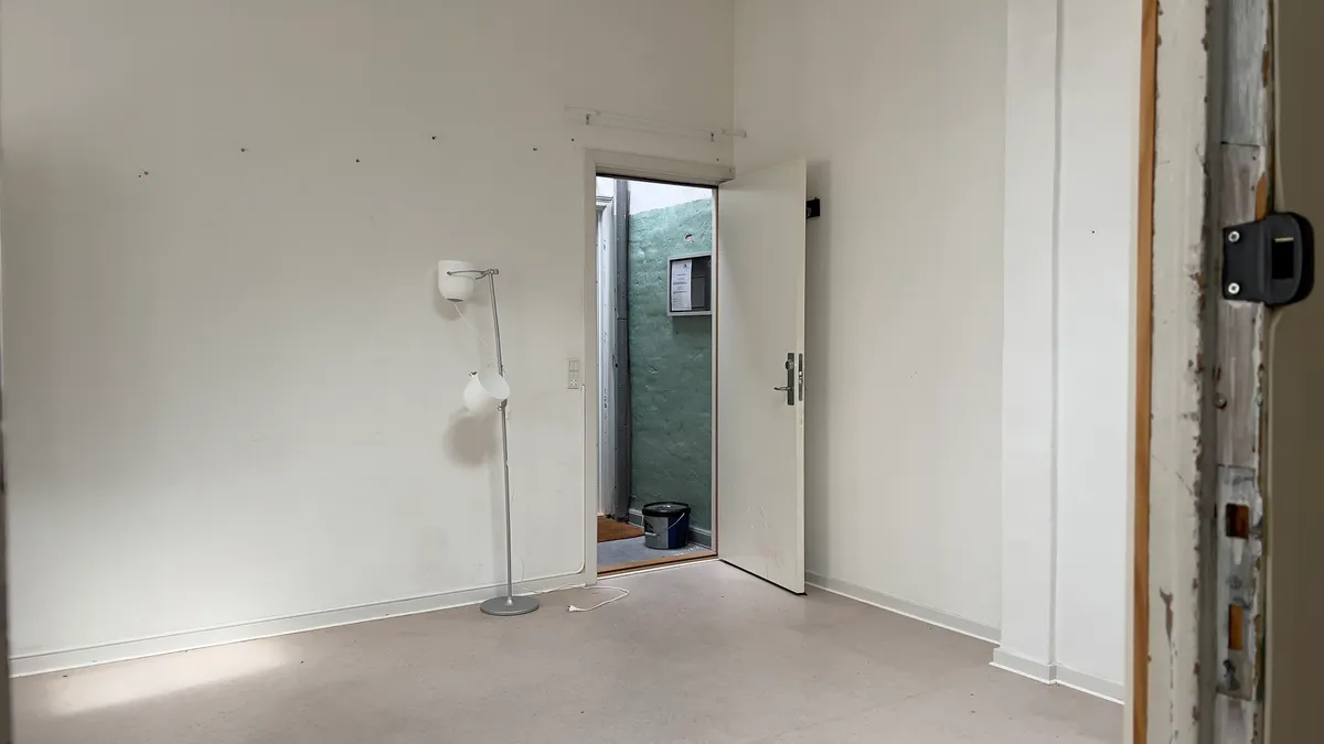 En tom stue med en hvid væg og et åbent hvidt dørblad, en lampe og en bøtte