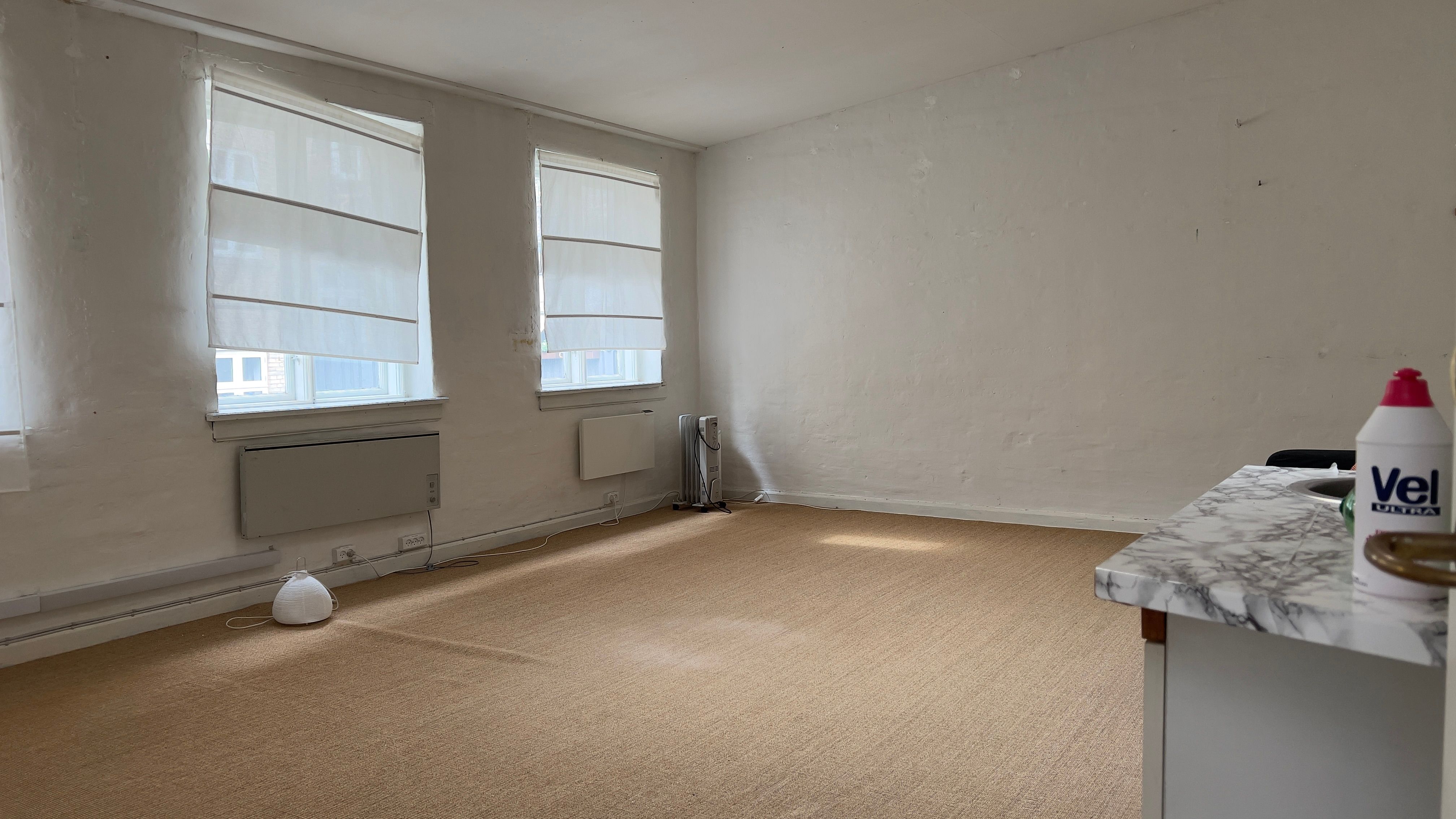 En tom kontorkontorrum med hvide vægge, to vinduer med hvide gardiner, og en marmorbord med en sprayflaske fra Vel Ultra på