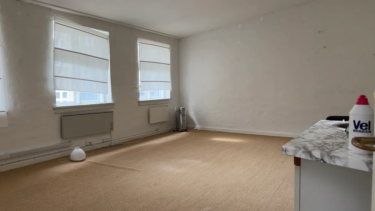 En tom kontorkontorrum med hvide vægge, to vinduer med hvide gardiner, og en marmorbord med en sprayflaske fra Vel Ultra på