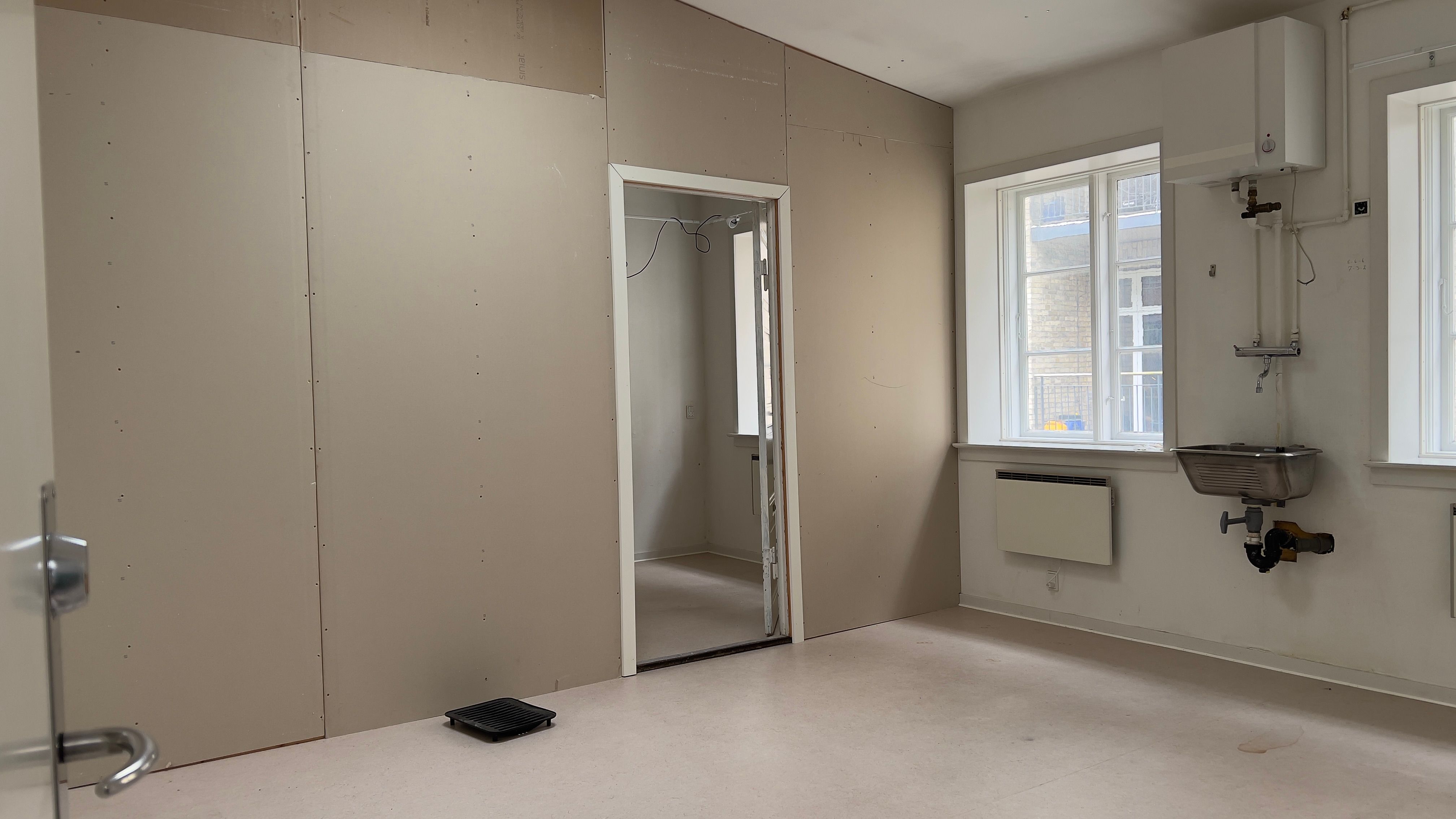 En tom kontorrum med lyse tavle og skraldespand ved døren og et vindues med udsigt til bygning og et skur