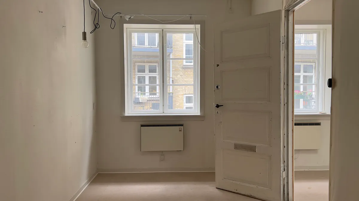 Rummet med hvide vægge og lysegult gulv har et stort hvidt dørblad og to små vinduer med udsigt til en bygning med en balkon