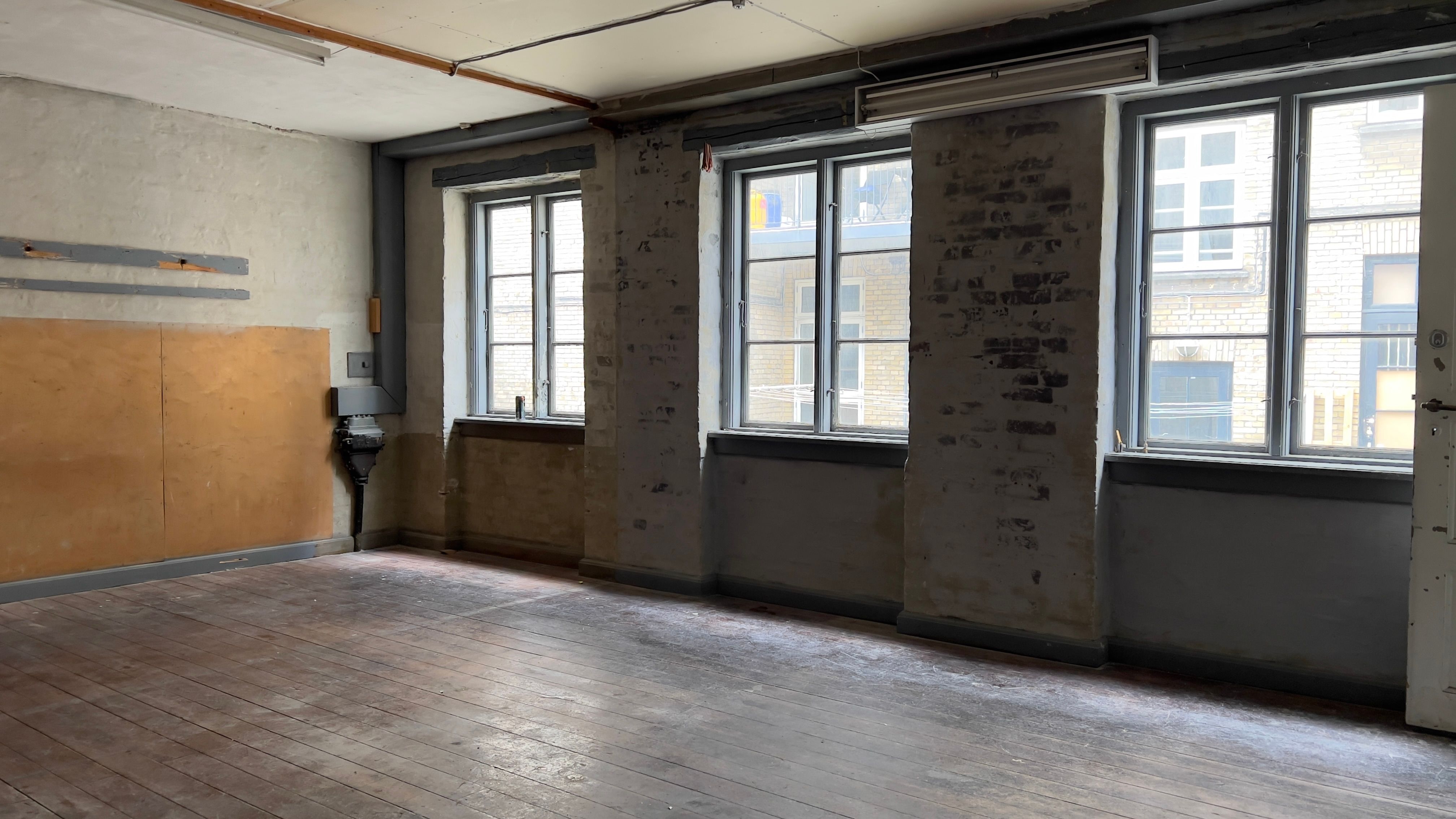 Et stort, tomt rum med lyse, trægulve og flere vinduer, som udmønter i samme stilart som de lyseblå døre, som er synlige