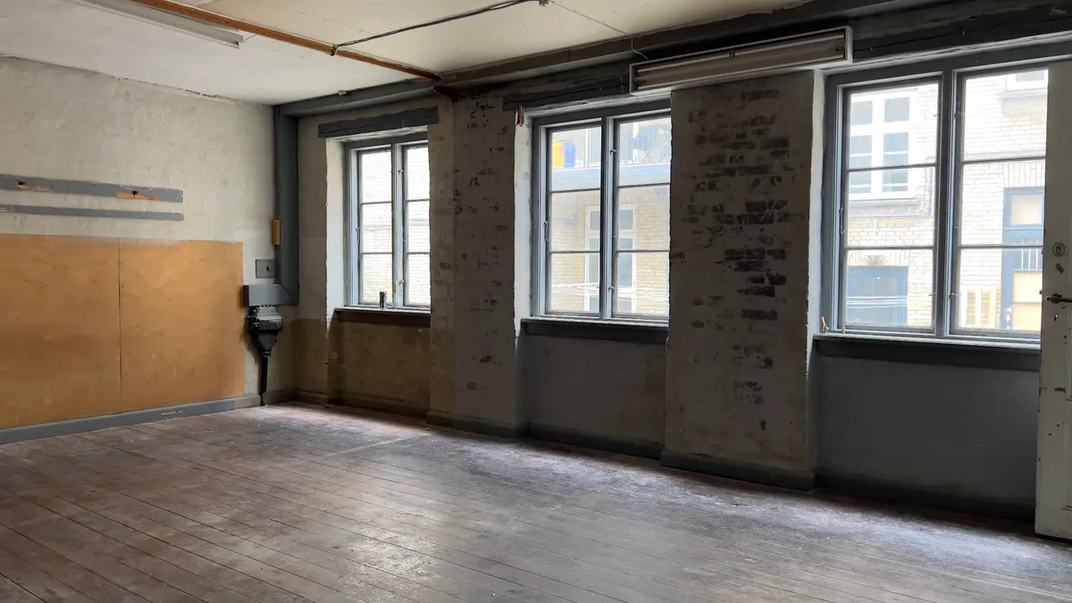 Et stort, tomt rum med lyse, trægulve og flere vinduer, som udmønter i samme stilart som de lyseblå døre, som er synlige