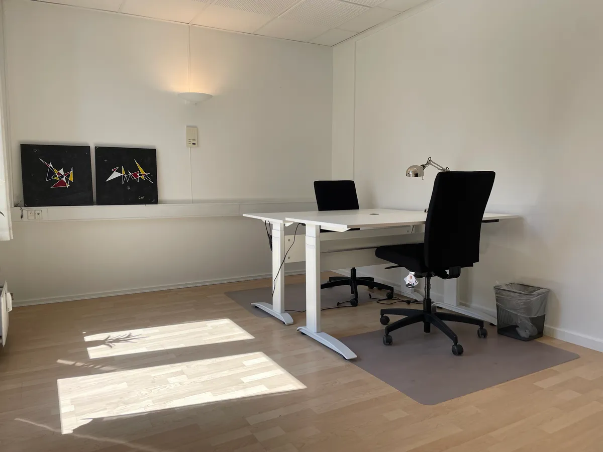 Kontor med to hvide hævesænkeborde og sorte kontorstole samt en lampe og to malerier på væggen.