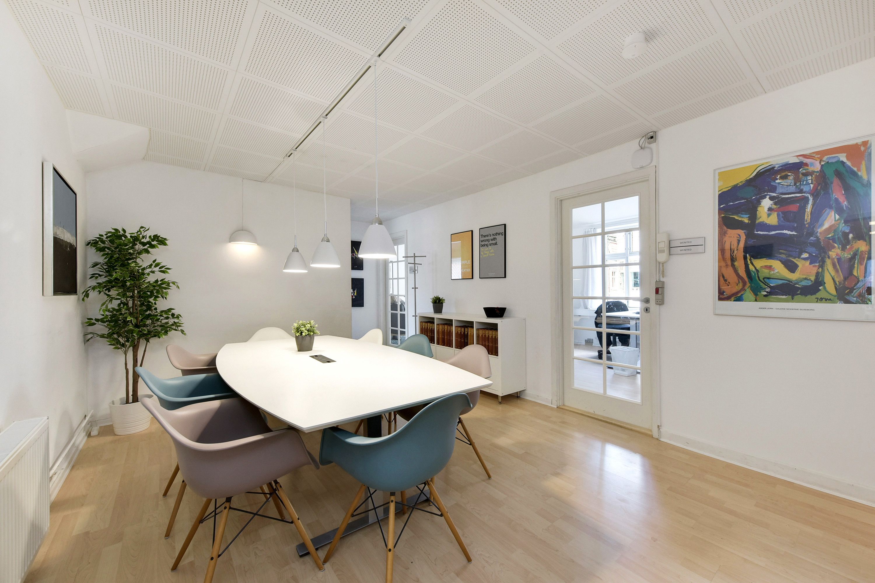 Hvidt kontorlokal med bord og stole, indrettet med hvide hævesænkeborde, plante og farverige vægmalerier
